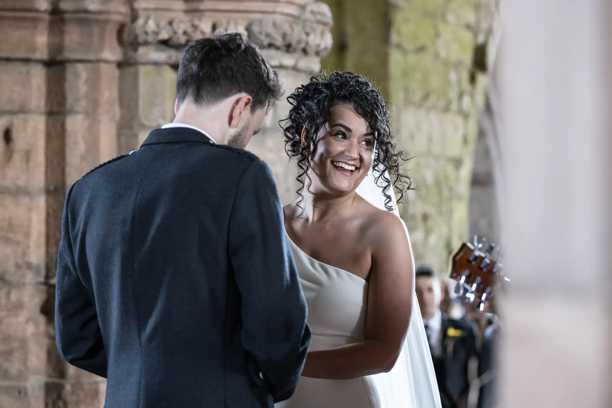 A couple stands together; the woman in a white dress smiles at the man in a dark suit while holding hands in a stone archway setting.