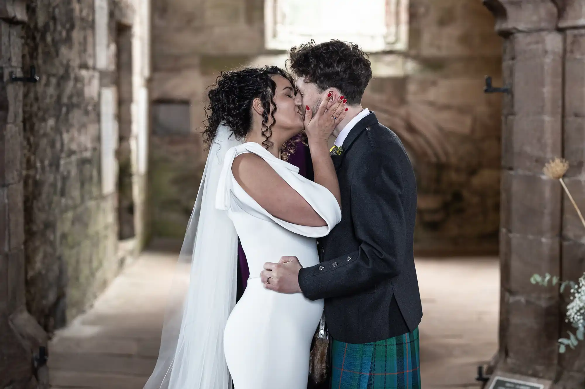 A couple kisses while getting married inside a historic stone building. The bride wears a white dress with a veil, and the groom wears a black jacket with a tartan kilt.