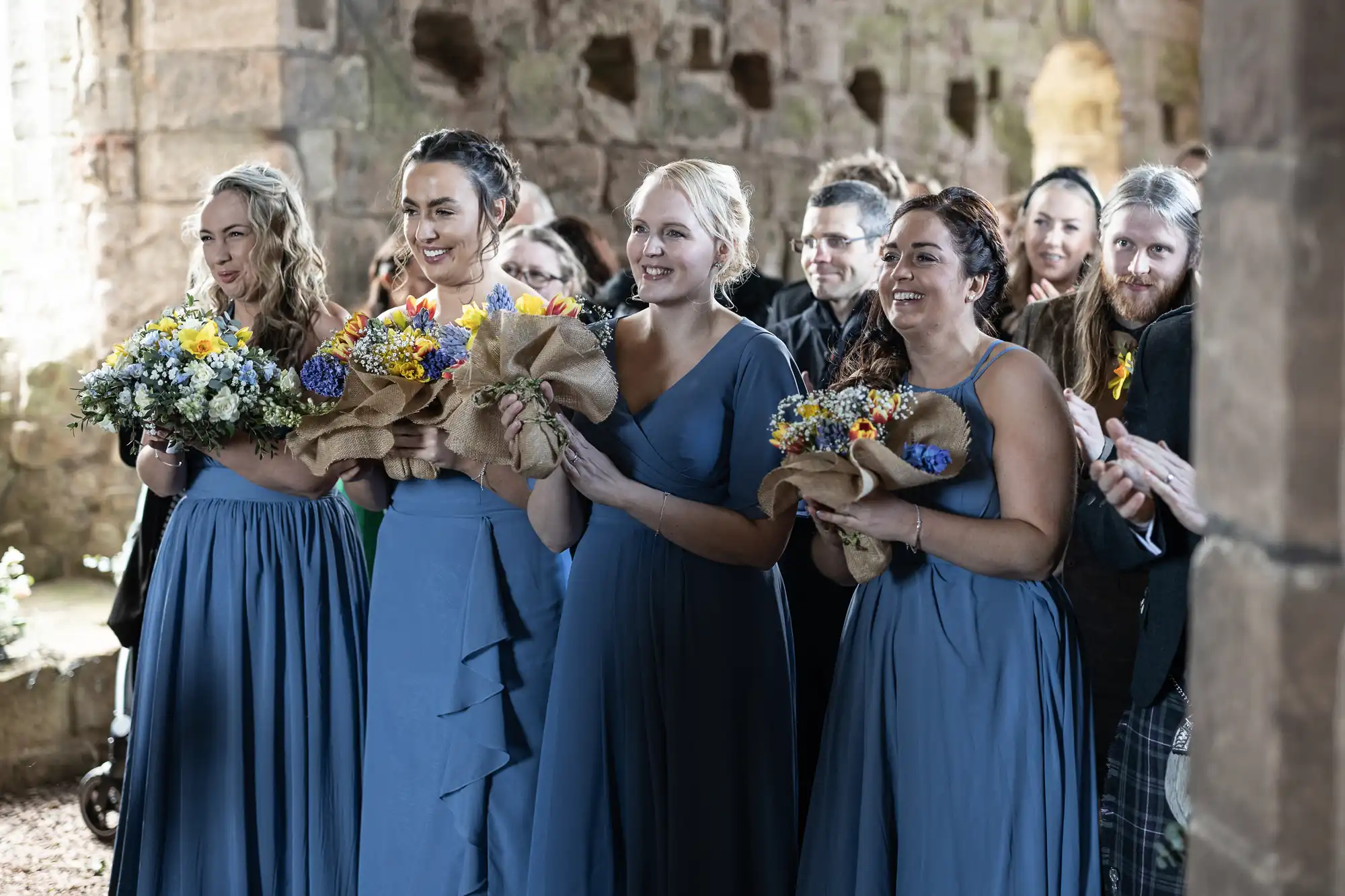 Four women wearing blue dresses holding bouquets, standing in a stone-walled indoor setting, with smiling people in the background.