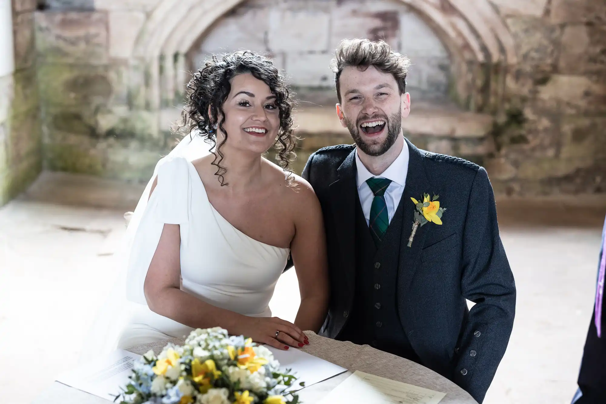 A bride in a white dress and a groom in a suit with a yellow boutonniere sit together at a table with a bouquet, both smiling.