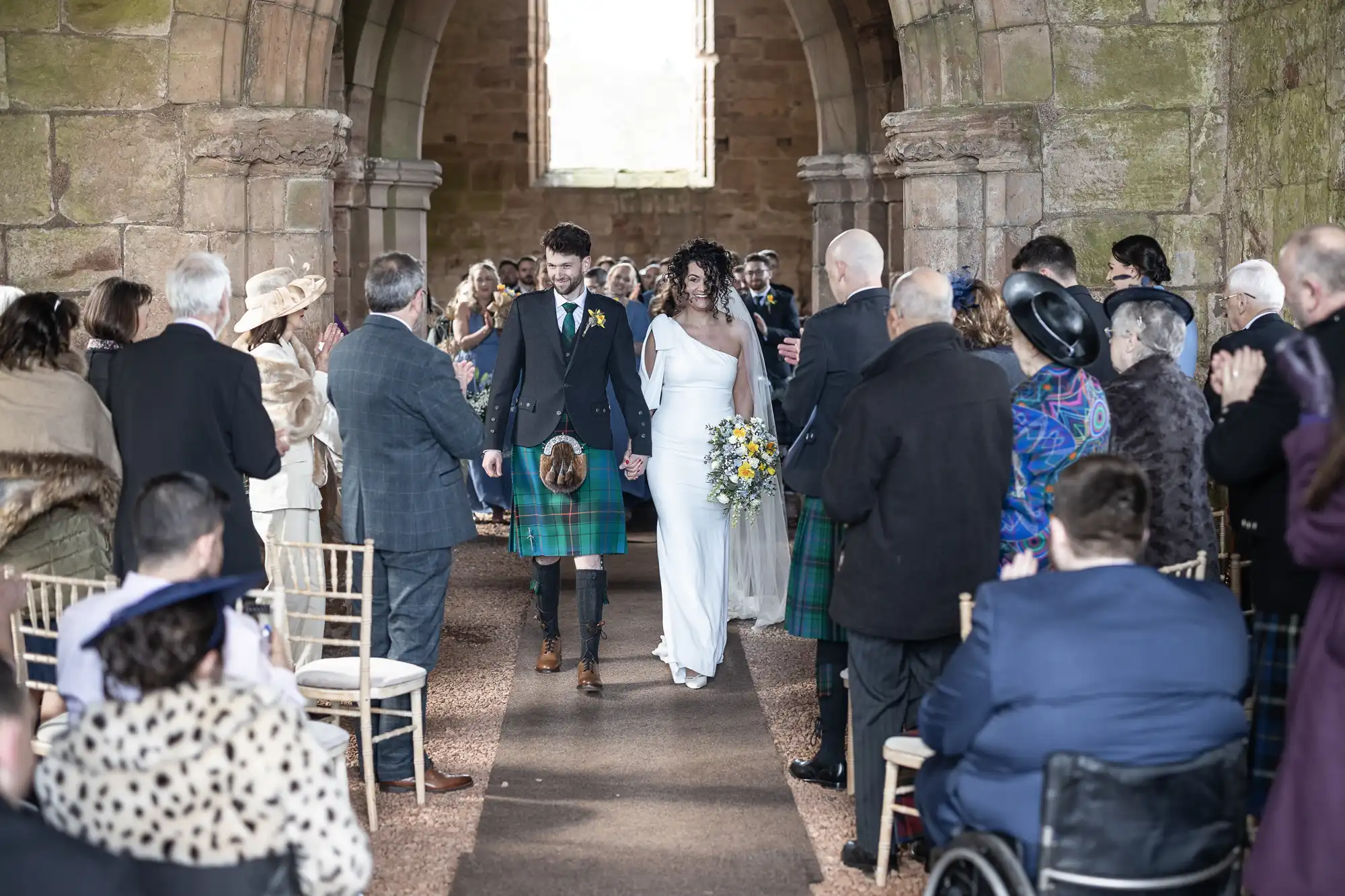 A couple, wearing traditional Scottish attire, walks down the aisle in an ancient stone church, surrounded by applauding guests.