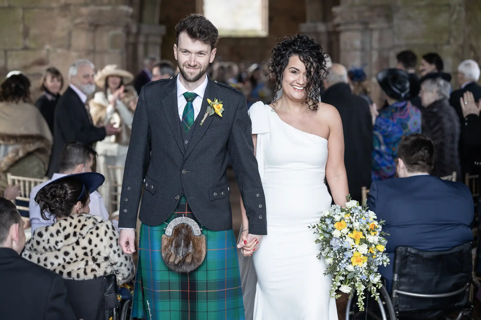 A bride in a white dress and a groom in a kilt hold hands and smile as they walk down the aisle of a stone building, with guests in the background.