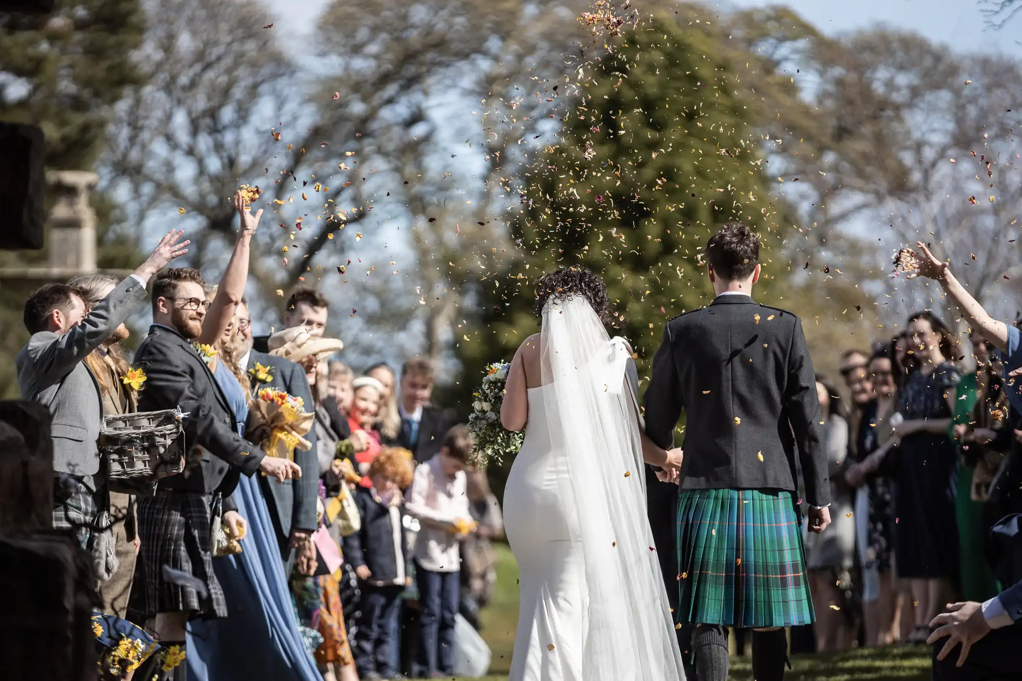 A newlywed couple walks down the aisle while guests toss flower petals at them. The groom is in a kilt, and the bride wears a white dress and veil. The outdoor setting features trees in the background.