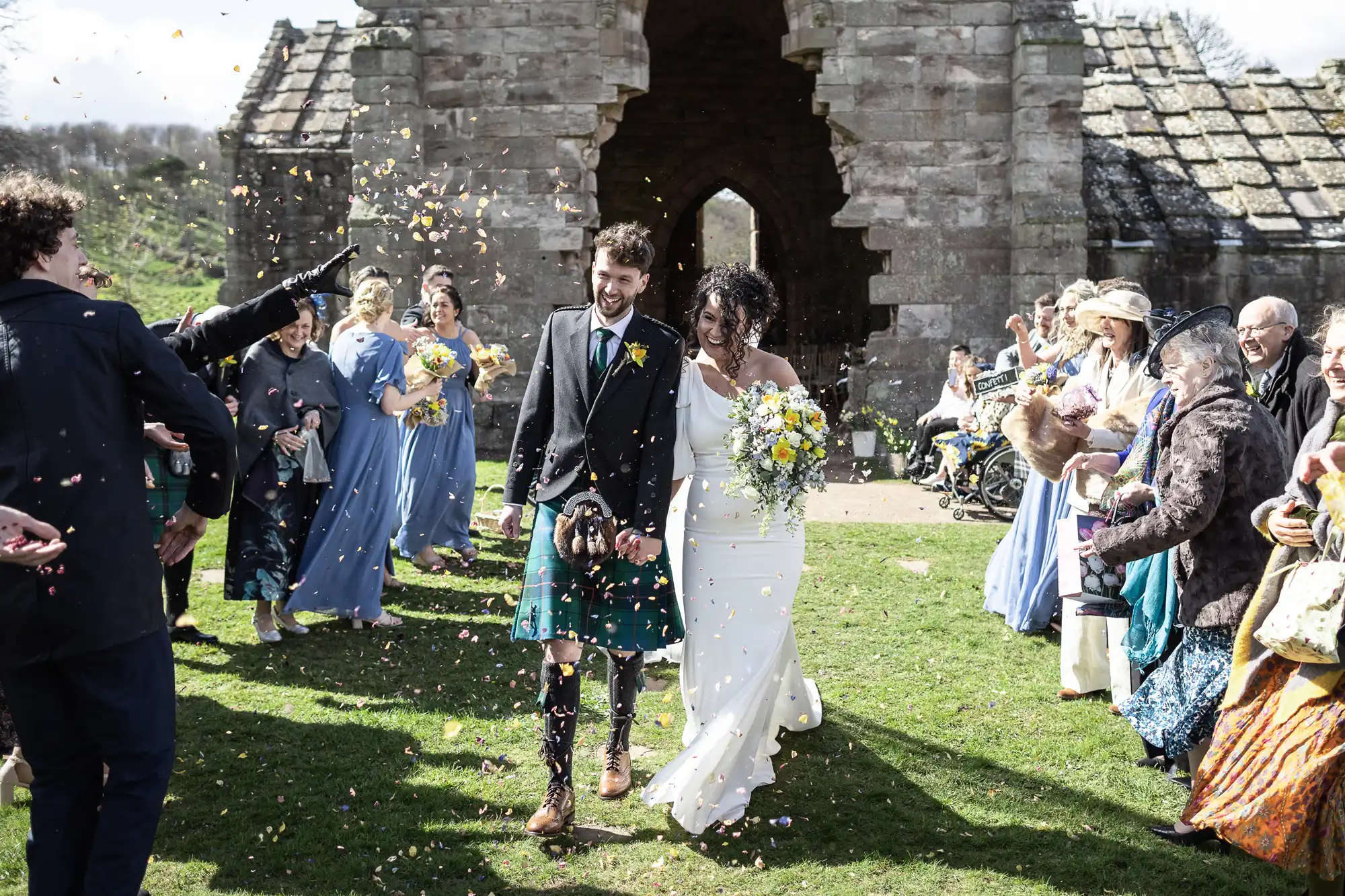 A couple in wedding attire walks down an aisle outdoors as guests shower them with flower petals. The groom wears a kilt, and the bride holds a bouquet of flowers. Guests are dressed in colorful outfits.