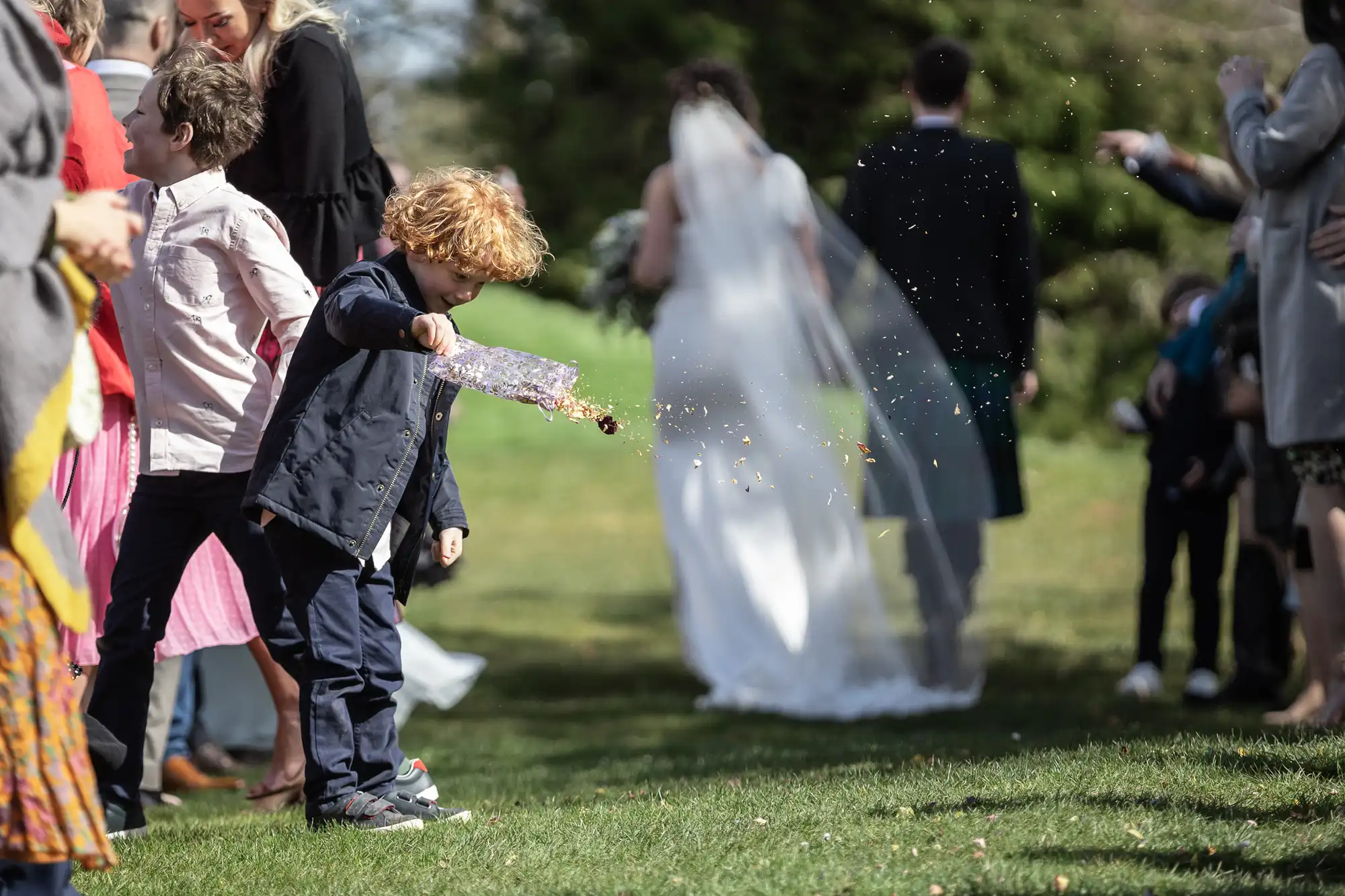 A young boy in a dark suit and blue sneakers is tossing flower petals on a grassy path as a bride and groom, along with other guests, walk forward.