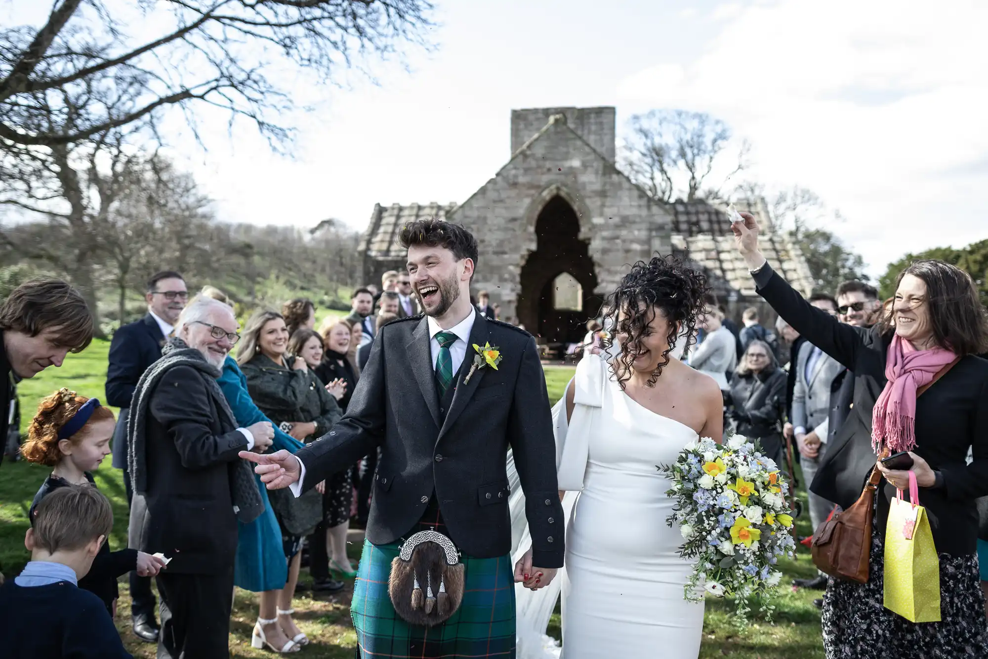 A joyful couple is walking down the aisle outdoors after a wedding ceremony, surrounded by smiling guests, with an old stone structure in the background.