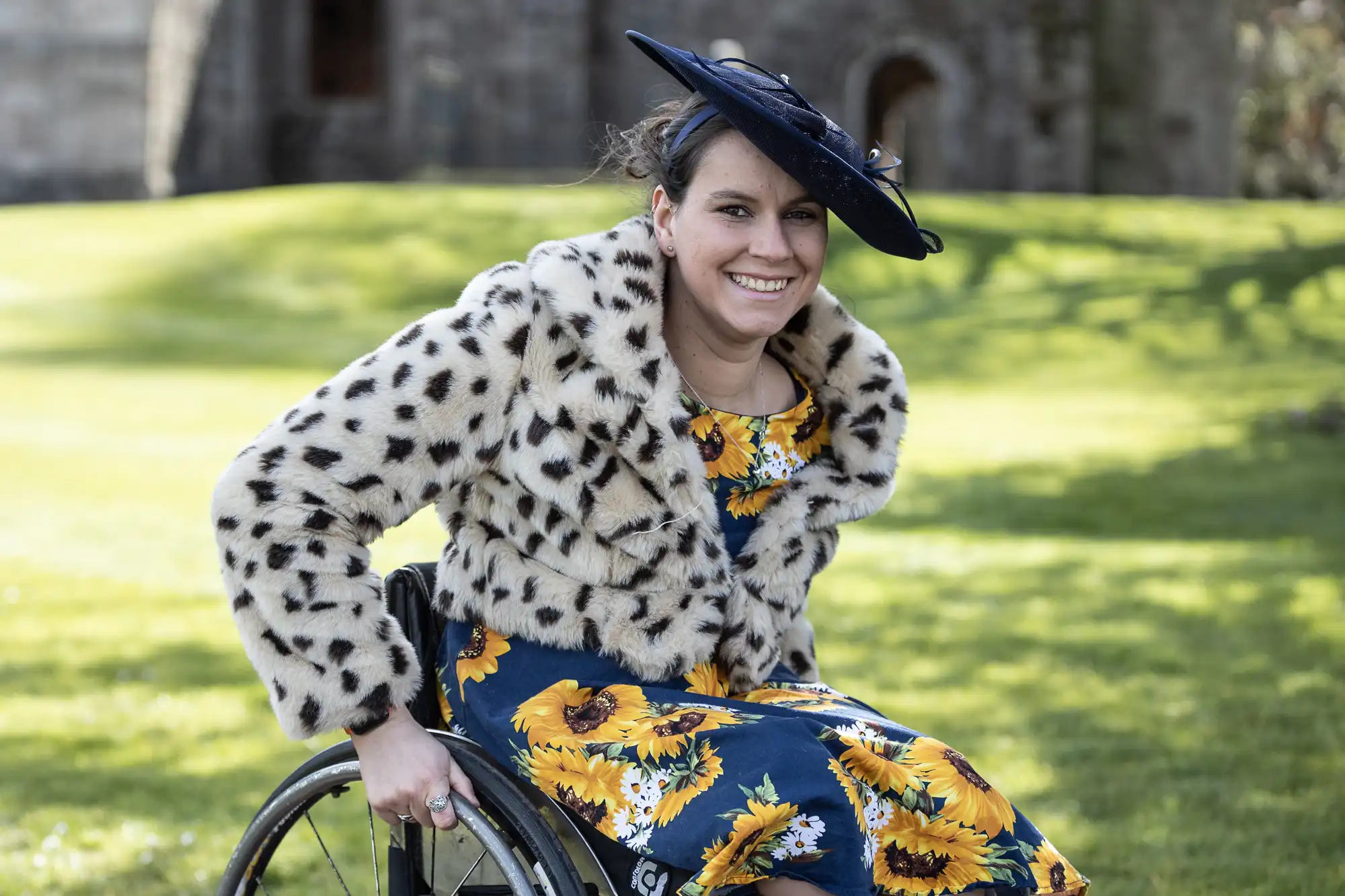 Smiling woman in a wheelchair, wearing a leopard print coat, sunflower-patterned dress, and black hat, outdoors in a grassy area with a background of a stone building.