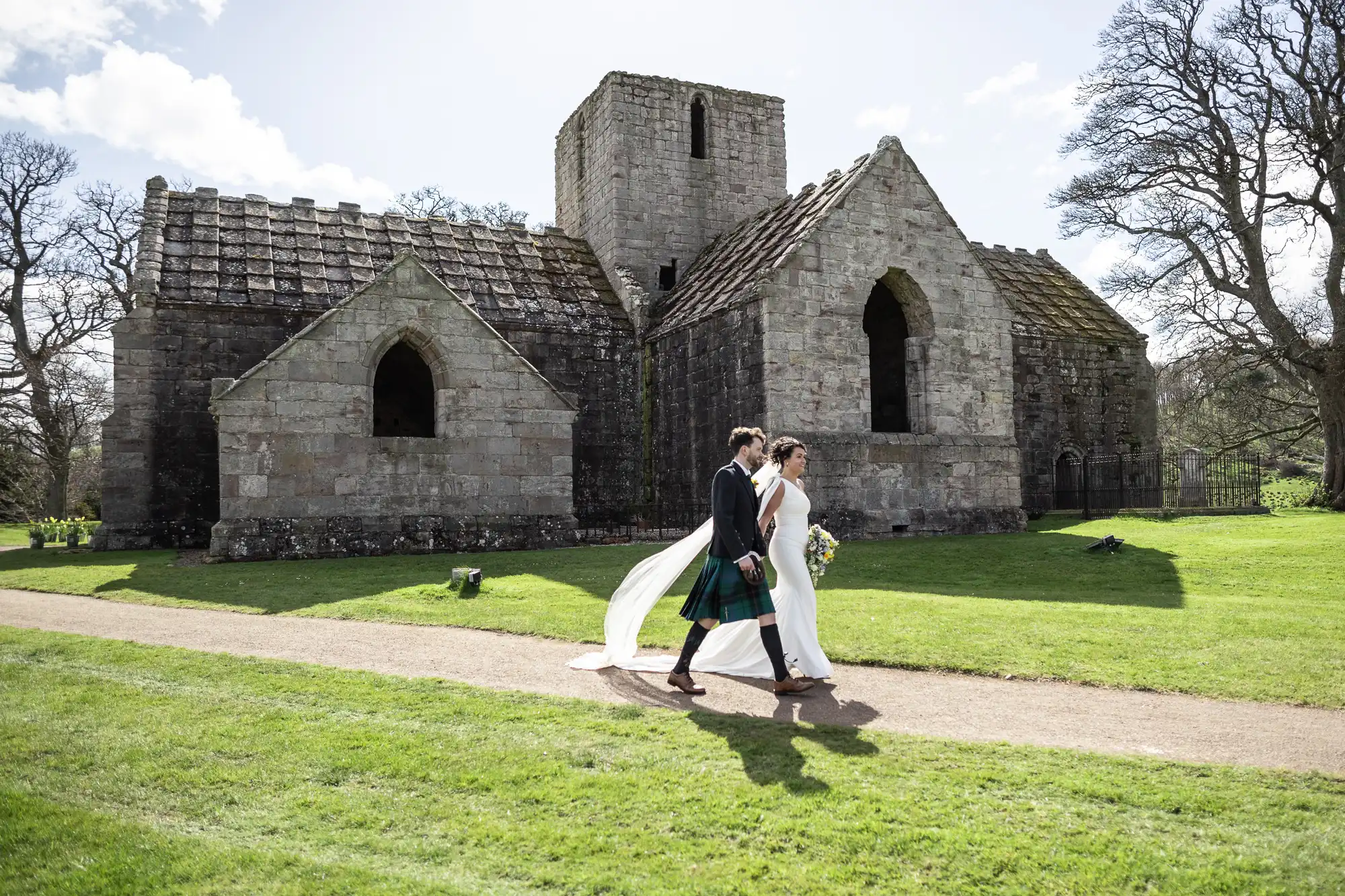 A couple, the groom in a kilt and the bride in a white dress, walk hand in hand on a path in front of an old stone church on a sunny day.