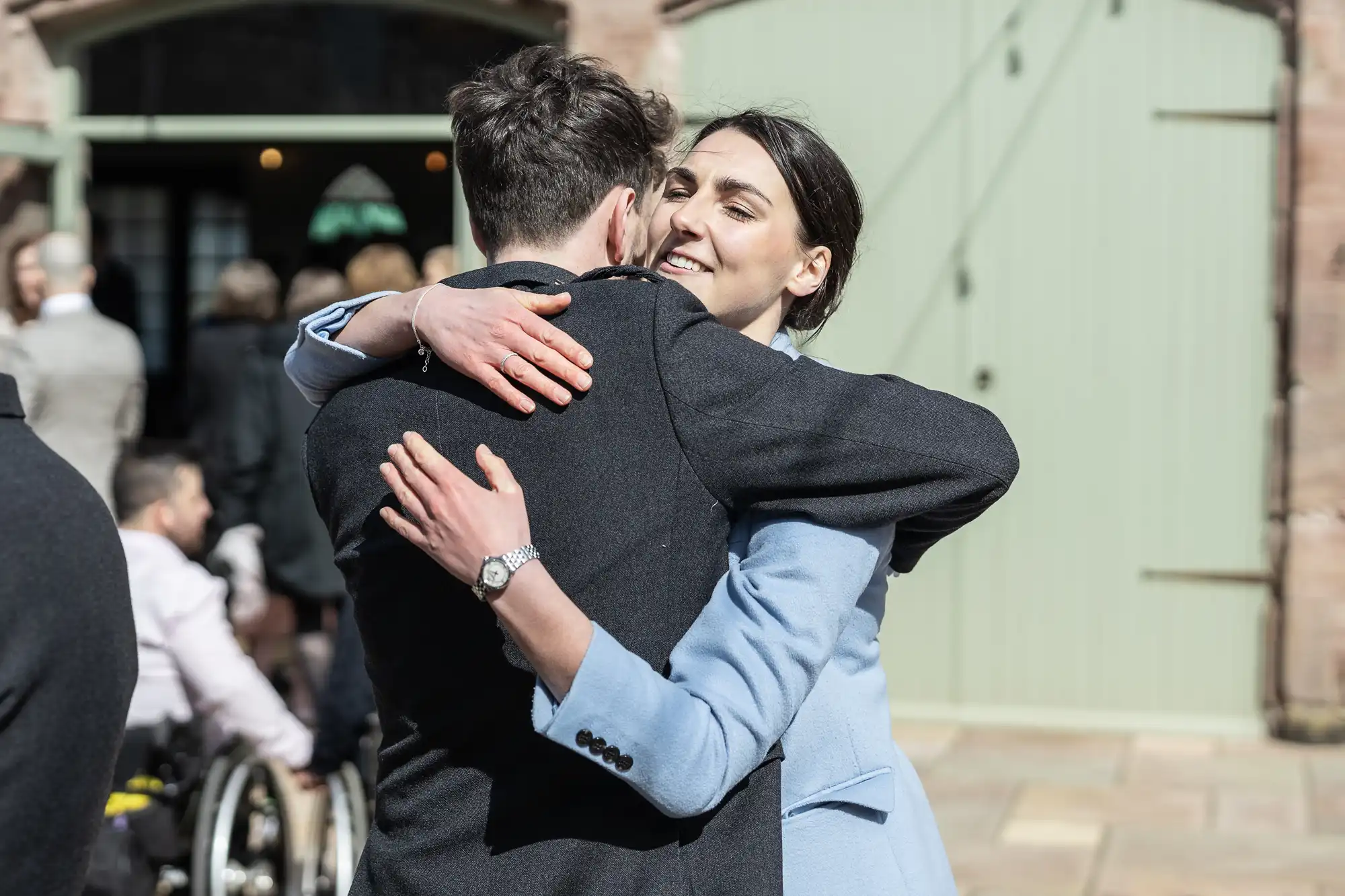 A man and woman embrace outdoors near a building with a green door, while others, including a person in a wheelchair, are visible in the background.