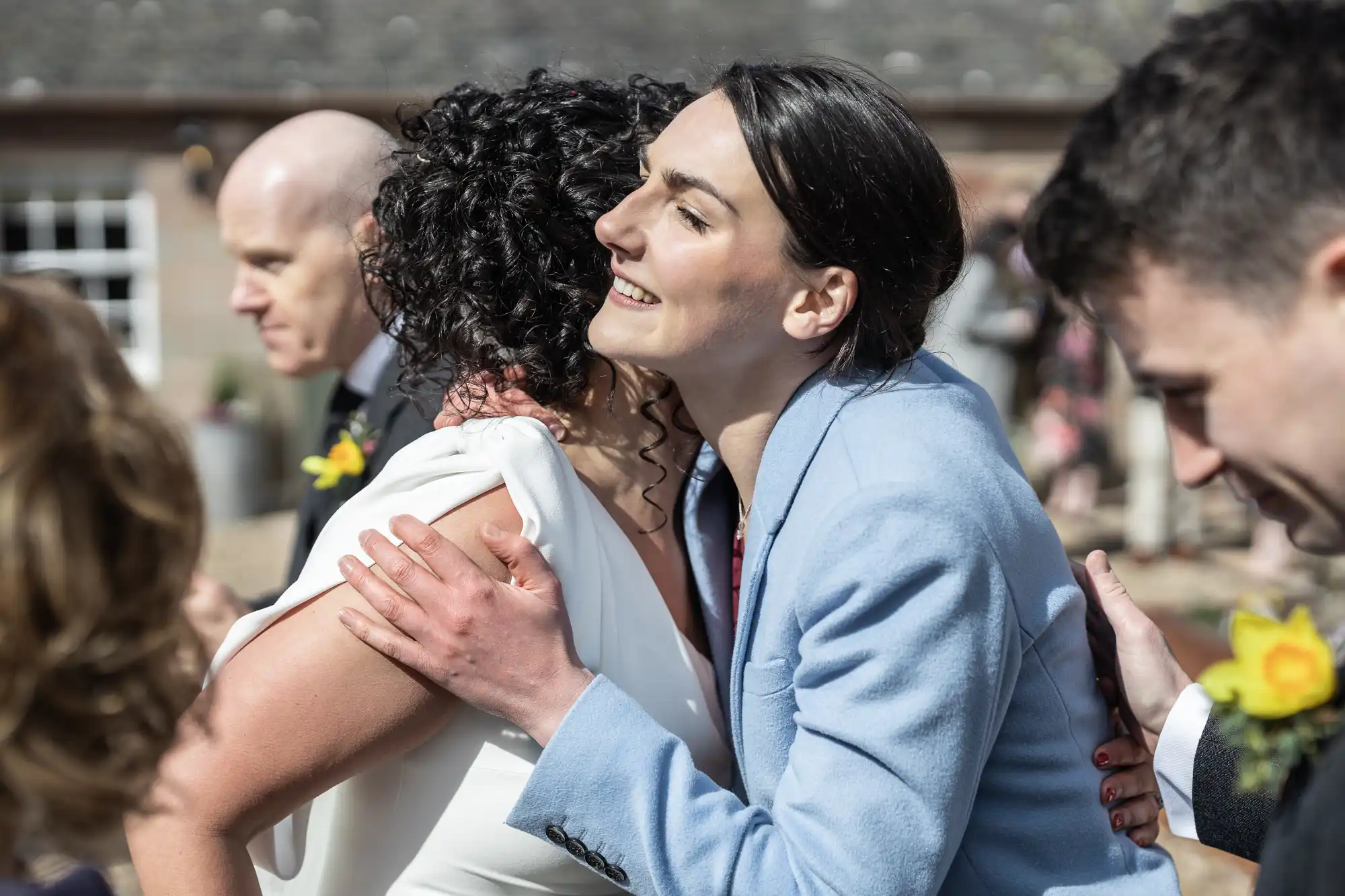 Two people embrace at an outdoor event; one person wears a white dress and the other is in a light blue suit. Other attendees can be seen in the background.