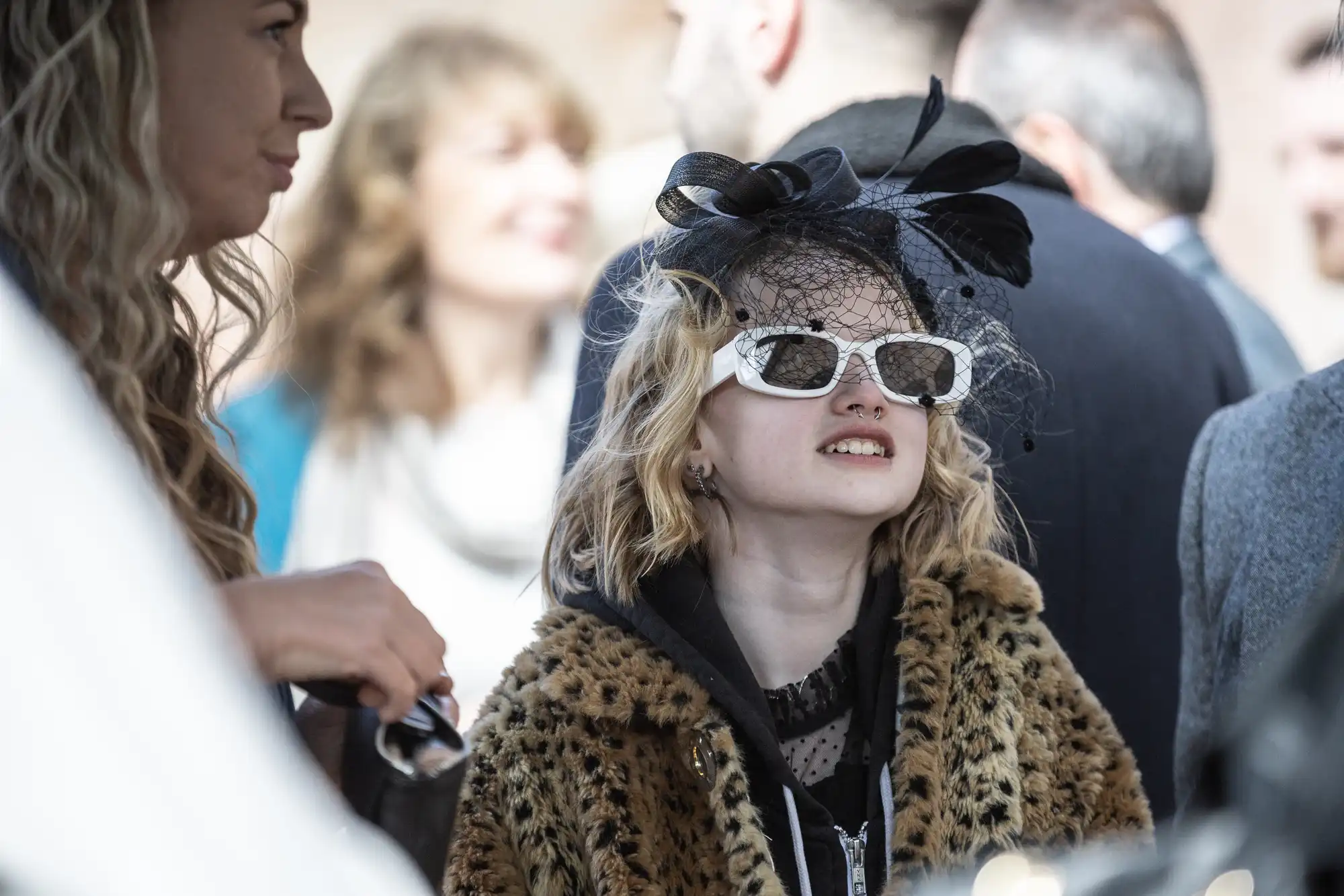 A person wearing a leopard print coat, white sunglasses, and a black fascinator stands outdoors among a group of people.