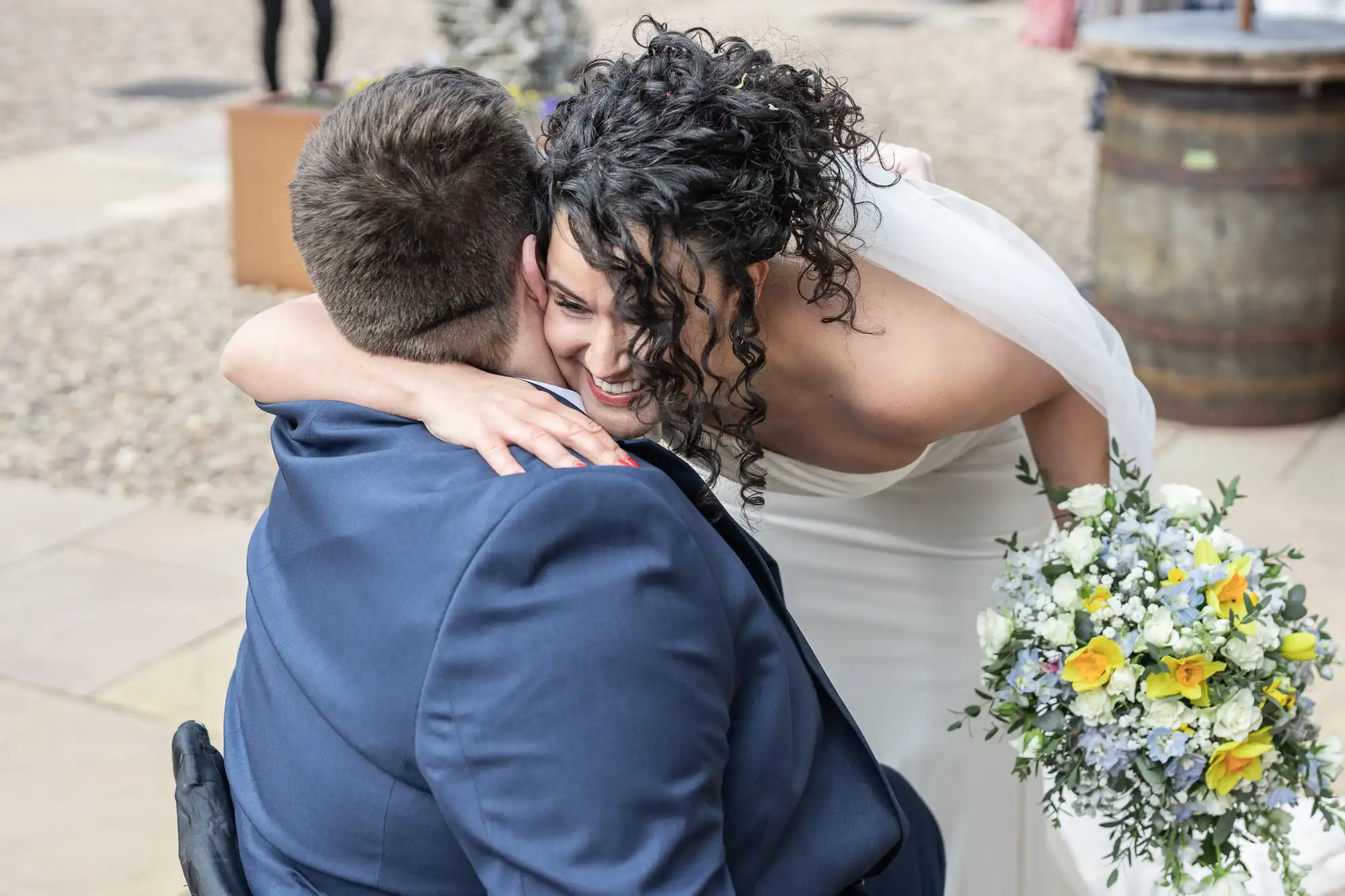 A bride in a white dress leans over to hug a person in a wheelchair, holding a bouquet of yellow, white, and blue flowers.