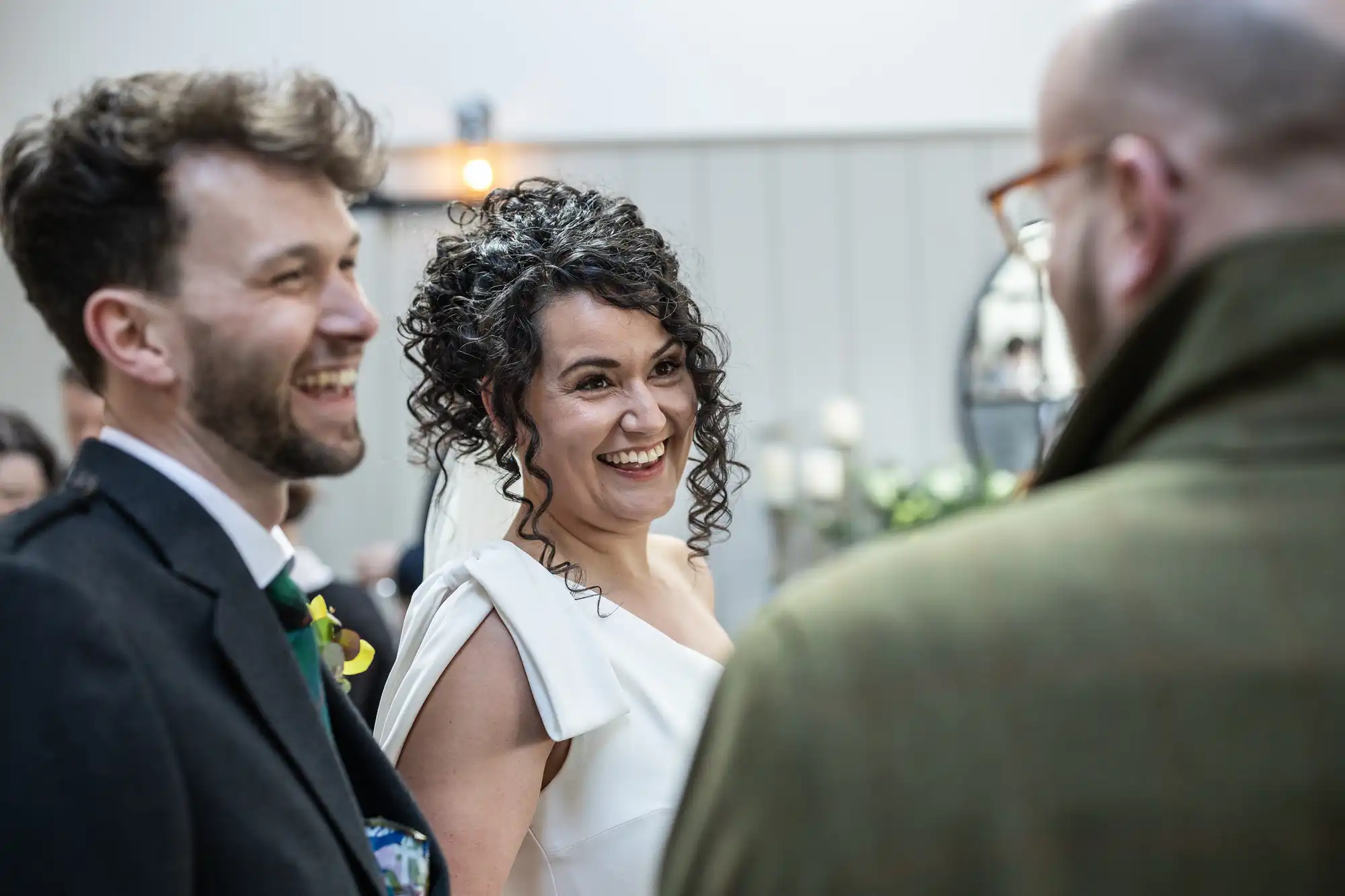 A bride and groom in formal attire are smiling and conversing with a guest. The setting appears to be an indoor venue.