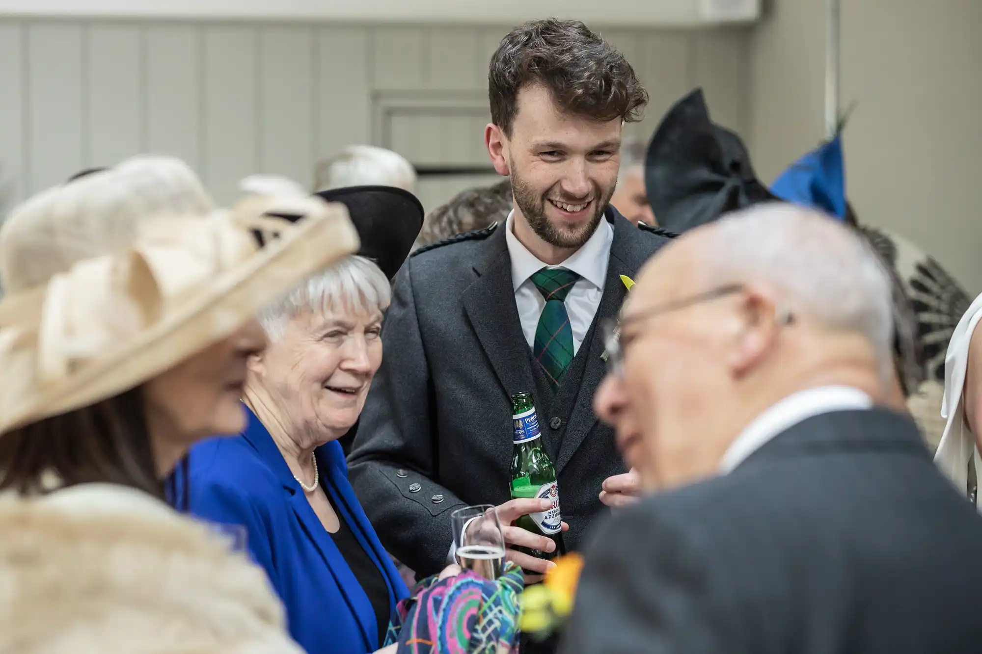 A man in a grey suit and tie holds a bottle of beer, smiling and conversing with three older individuals at a social event. The group is dressed formally, with one woman wearing a hat.