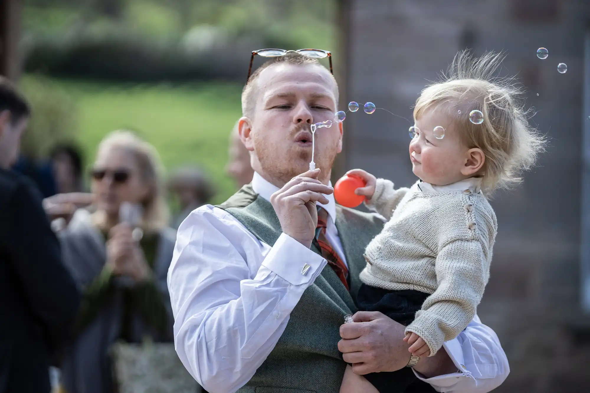 Man blowing bubbles while holding a toddler outdoors. The toddler, dressed in a knitted sweater, holds a red ball and looks at the bubbles. Other people are blurred in the background.