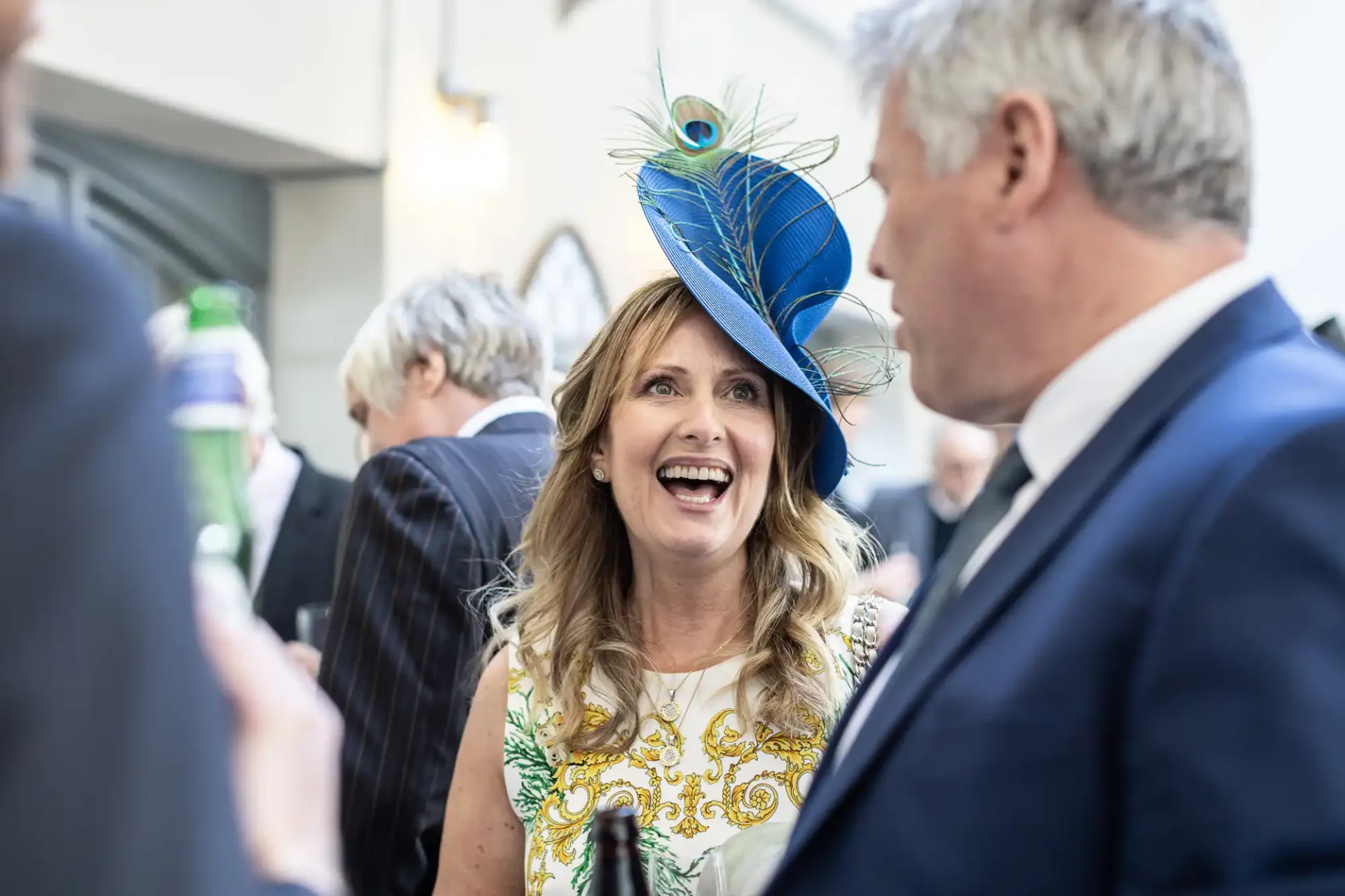 A woman wearing a decorative hat with a blue and green plume smiles while conversing with a man in a suit at a social event.