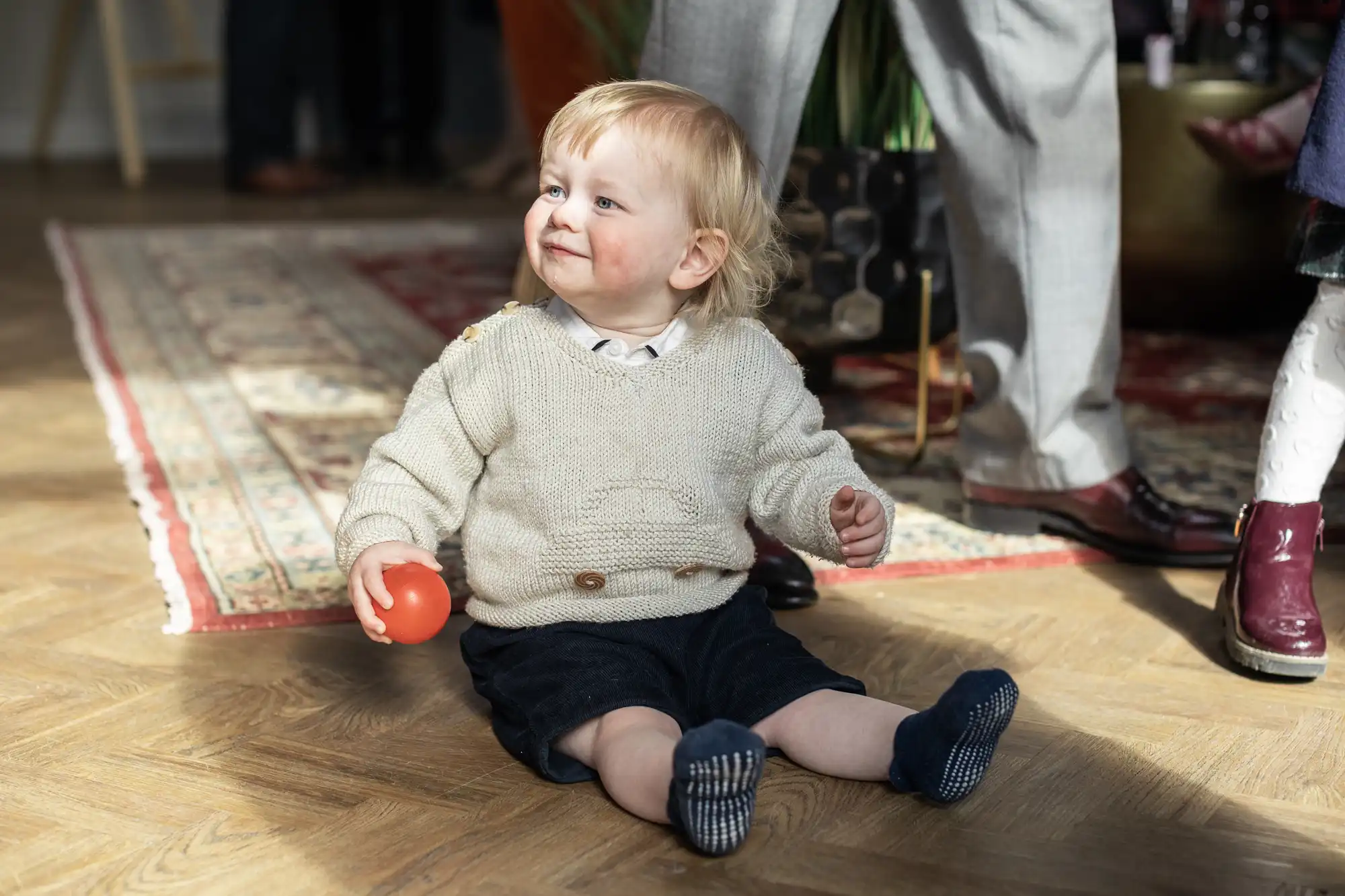 Young child with light hair wearing a cream sweater and dark shorts sits on a wooden floor holding a red ball. A patterned rug is in the background along with legs of adults standing nearby.