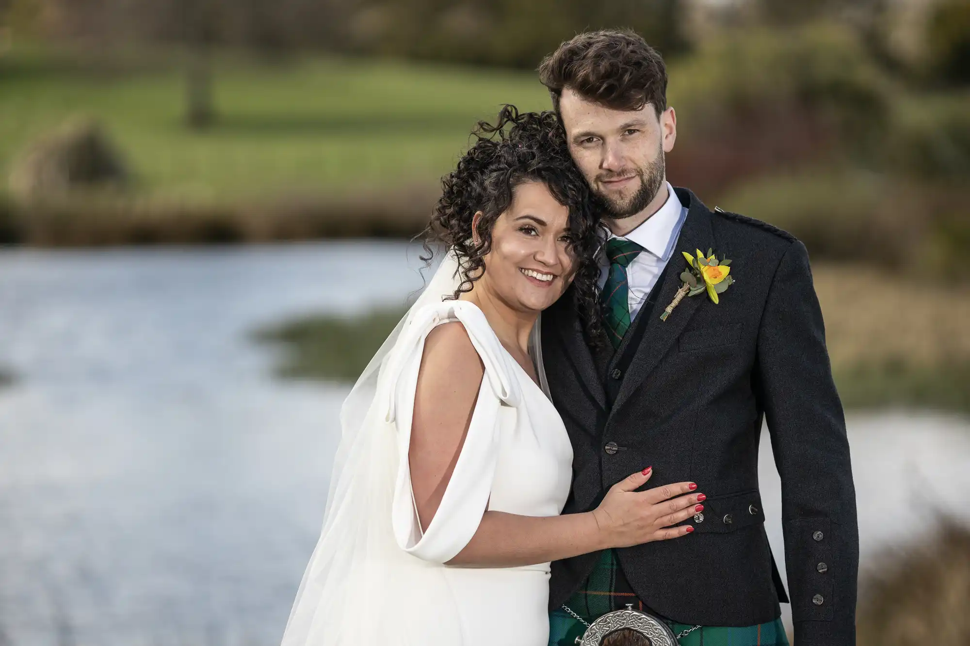 A couple stands by a body of water, the woman in a white dress and the man in a dark suit with a kilt, smiling at the camera.