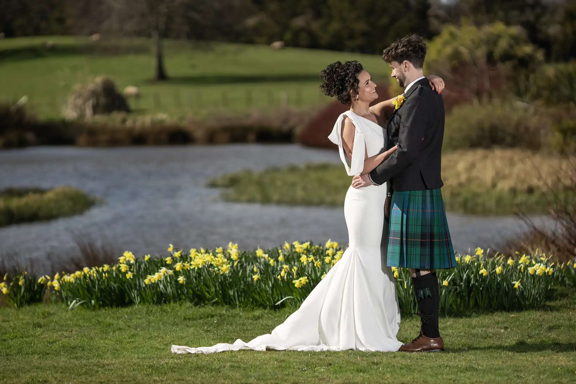 A bride in a white dress and groom in a kilt embrace beside a lake, with daffodils in the foreground and a green field in the background.