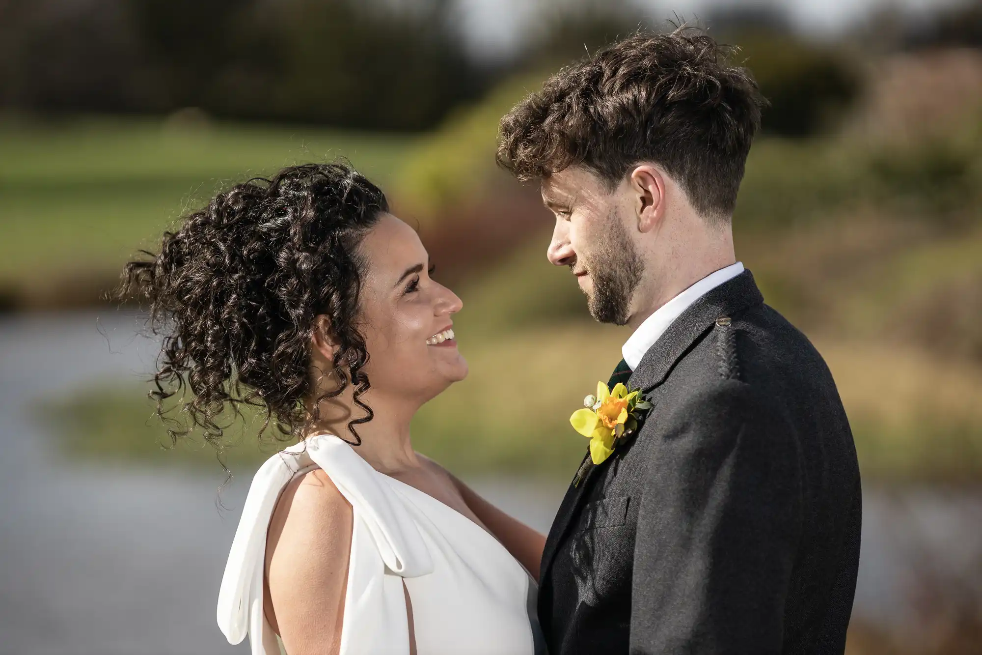 A couple in wedding attire looks at each other while standing outdoors. The woman has curly hair and wears a white dress; the man has short hair and wears a dark suit with a yellow flower boutonniere.