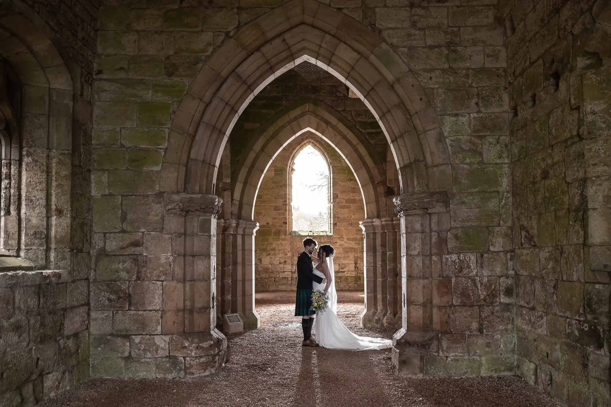 A couple stands in a stone archway, facing each other, in an ancient, stone-built setting, possibly a historical church or ruin. The bride is in a white gown, and the groom is in a dark suit with a kilt.