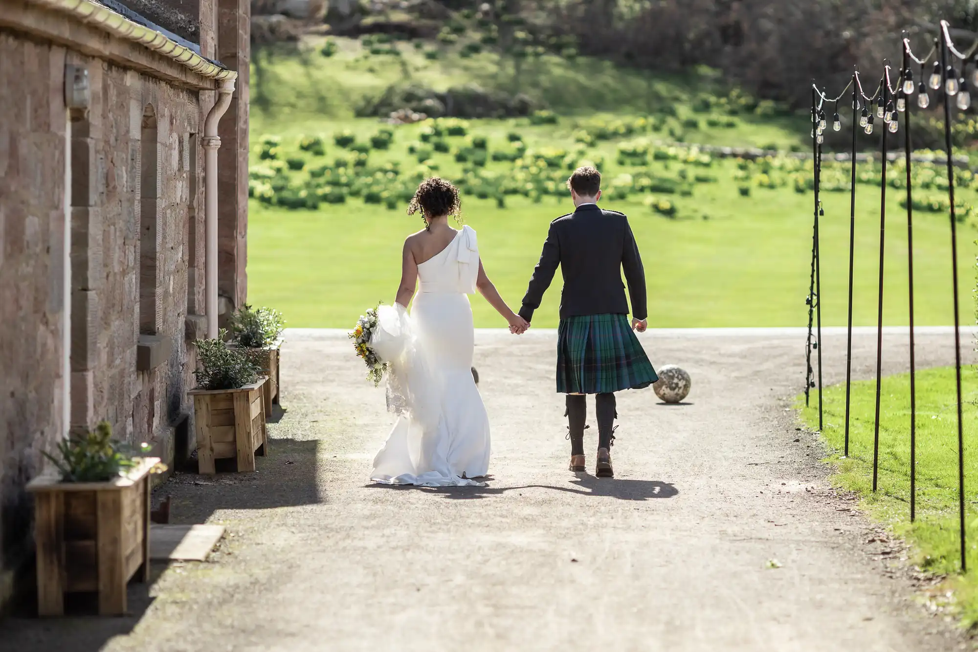 A bride in a white gown and a groom in a kilt hold hands and walk away from a stone building towards a green meadow.