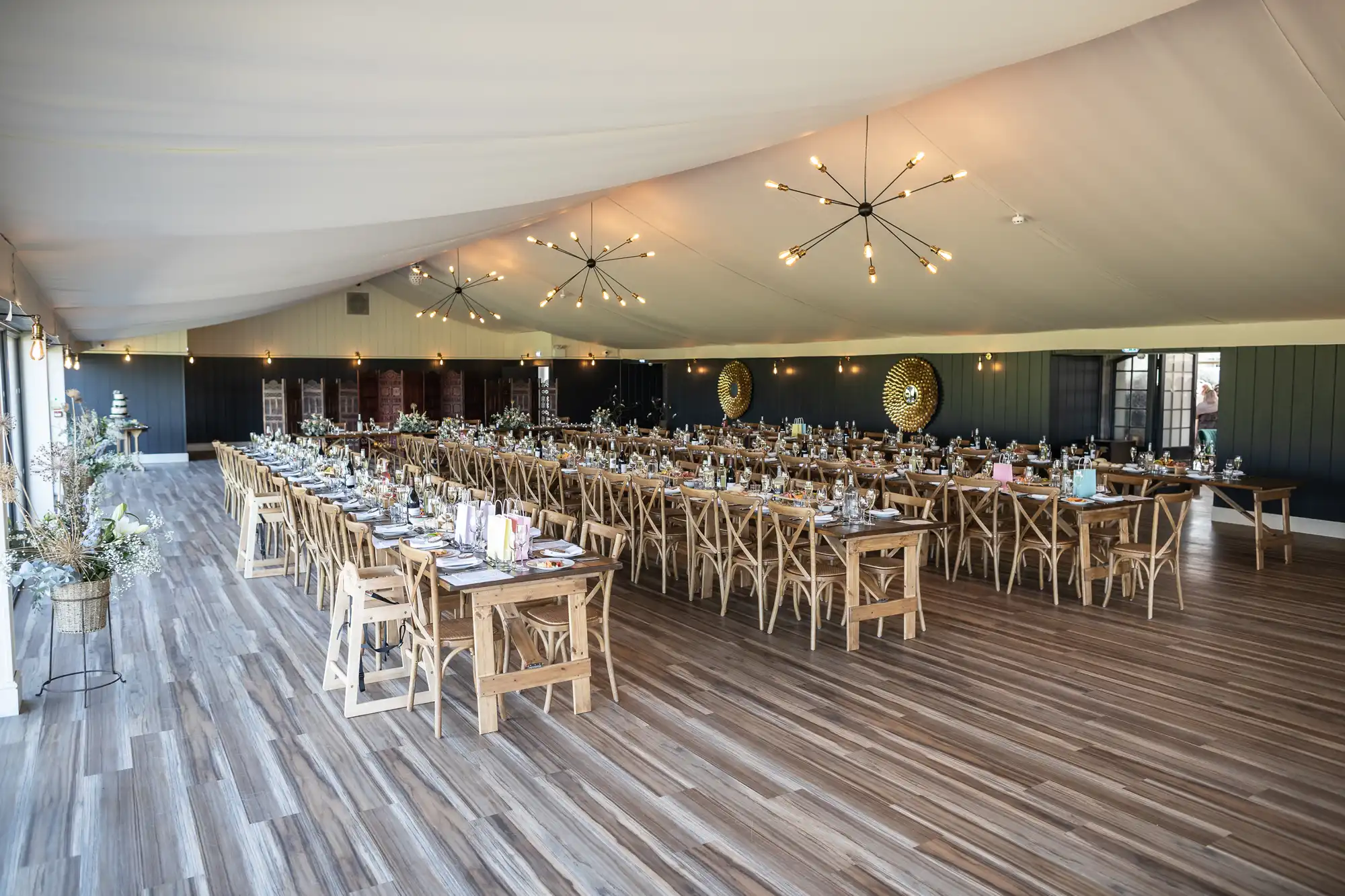 Large event space with wooden tables and chairs, set for a meal under a tented ceiling with modern chandeliers. Tables are arranged in rows, adorned with place settings and decorative centerpieces.