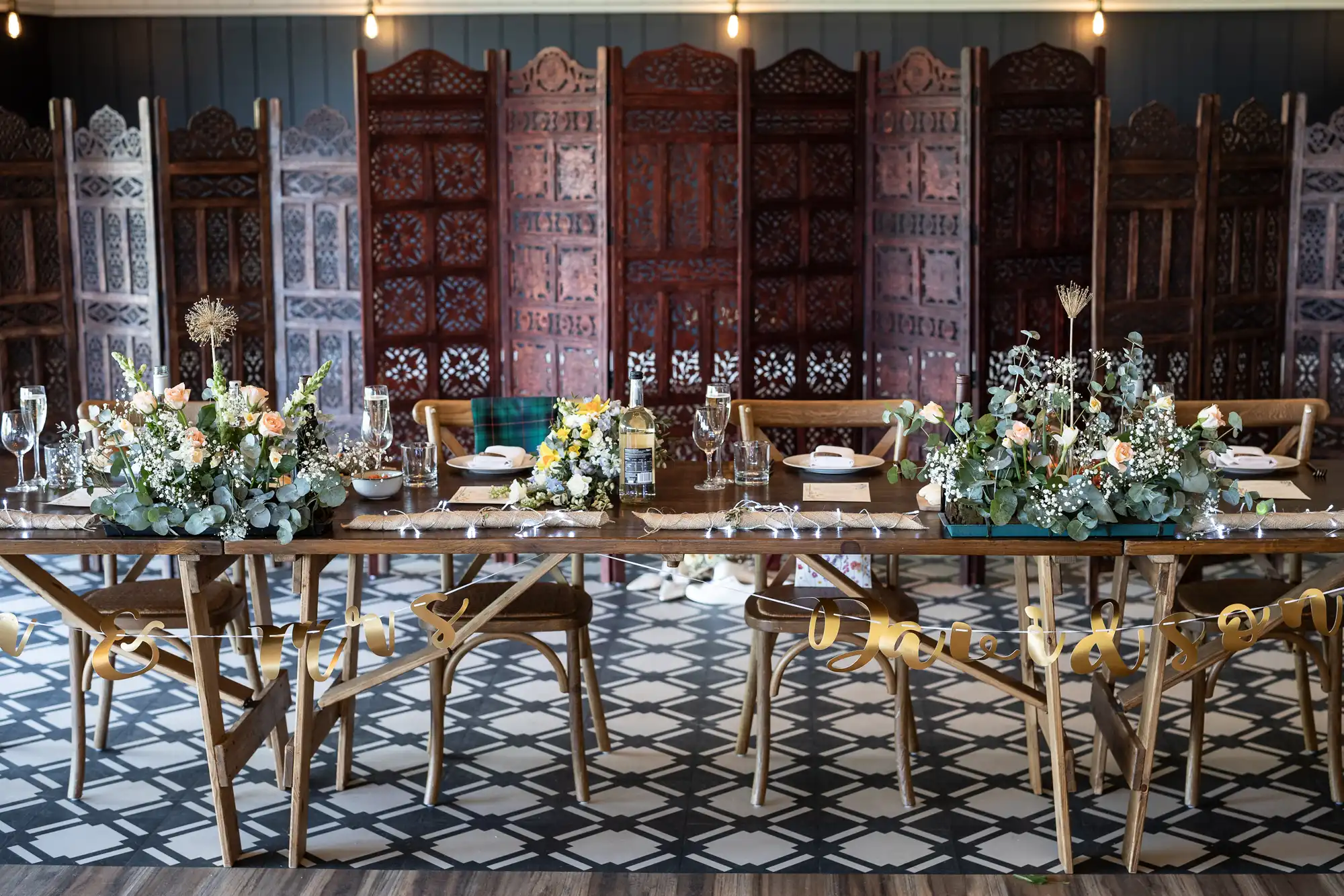 A decorated dining table set for an event, featuring floral centerpieces, glassware, and tableware, with wooden chairs and ornate room dividers in the background.