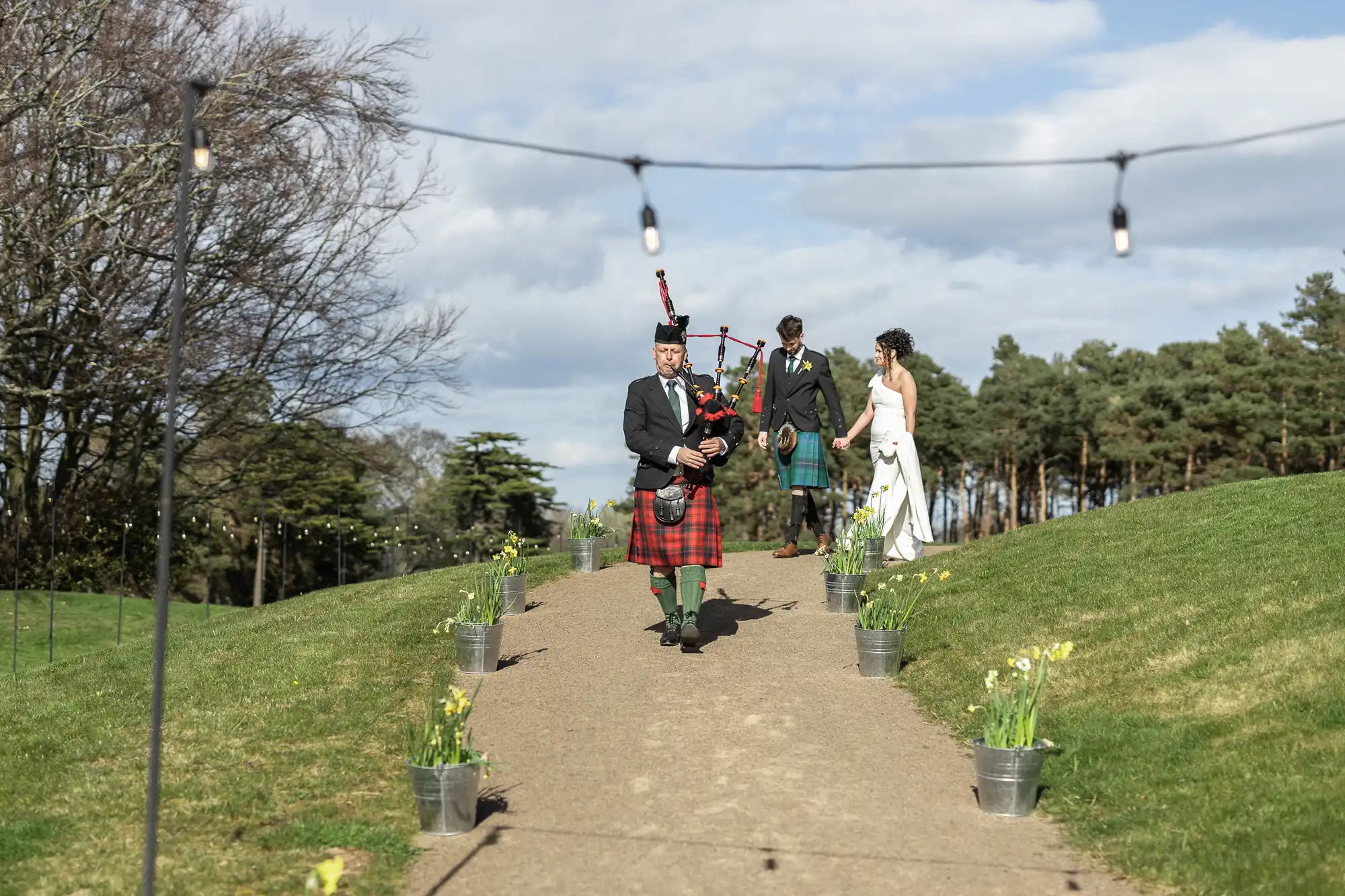 A bagpiper leads a wedding procession outdoors, followed by a groom and bride on a path lined with daffodils and string lights, with trees and a blue sky in the background.