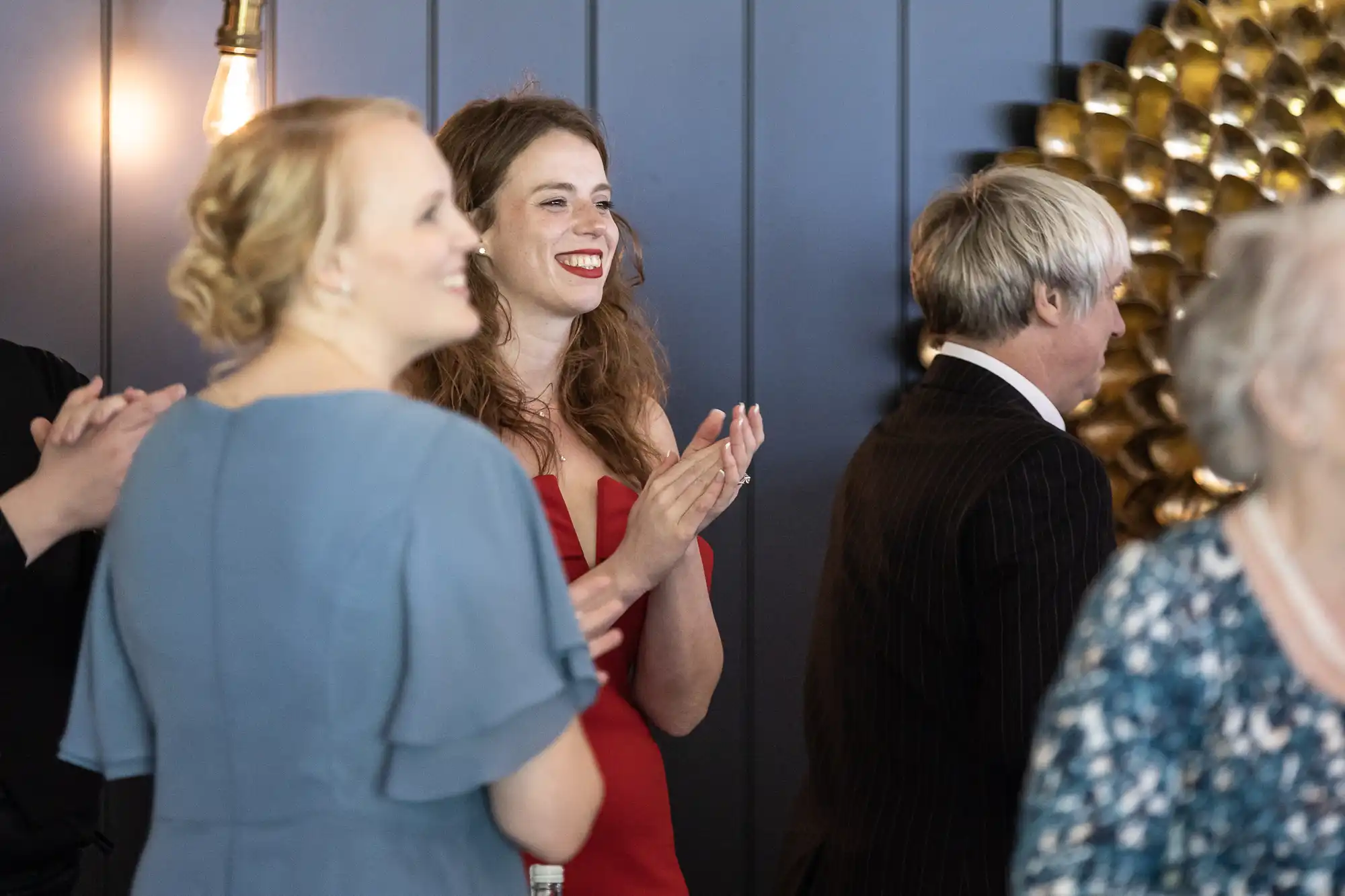 Group of people clapping, including women in blue and red dresses, and a person with short gray hair. They are in a room with blue paneled walls and a gold decorative object in the background.