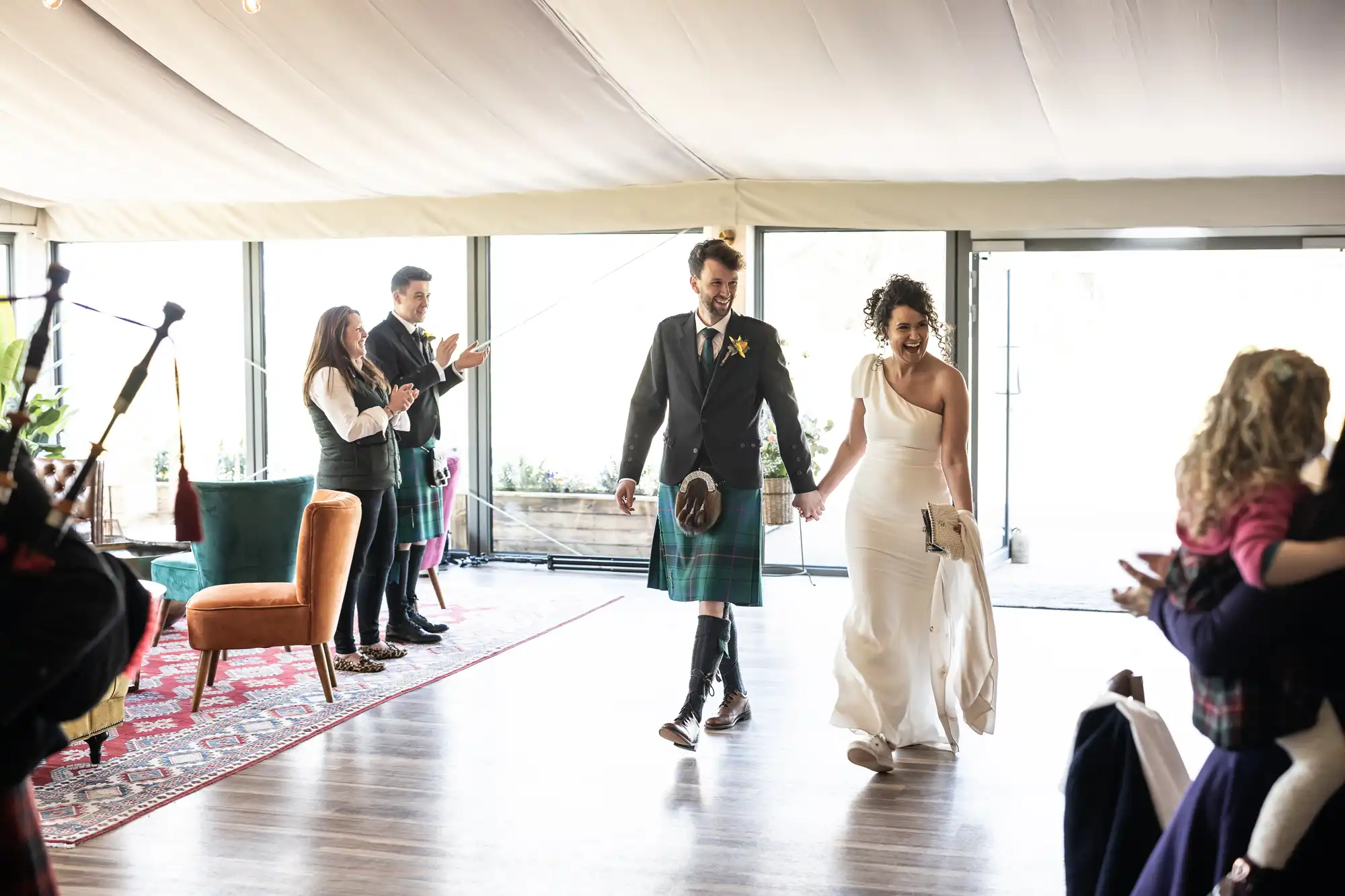 A couple, one in a white dress and the other in a kilt, walk hand in hand into a room as people stand and clap.