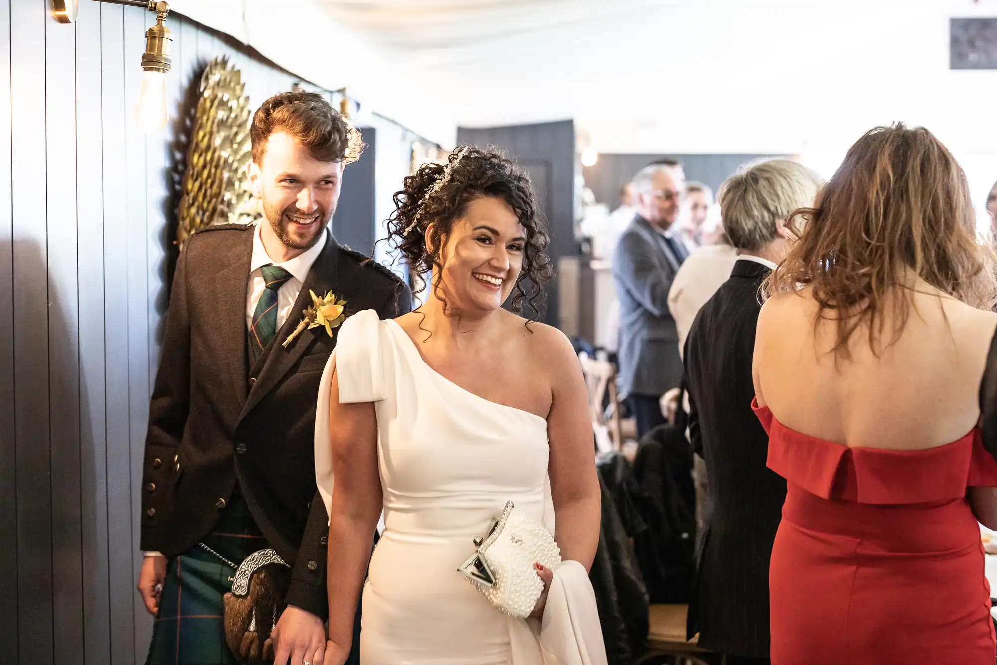 A couple dressed in formal attire, with the woman in a white one-shoulder dress and the man in a dark suit, stand together at a social event. Other guests are present in the background.