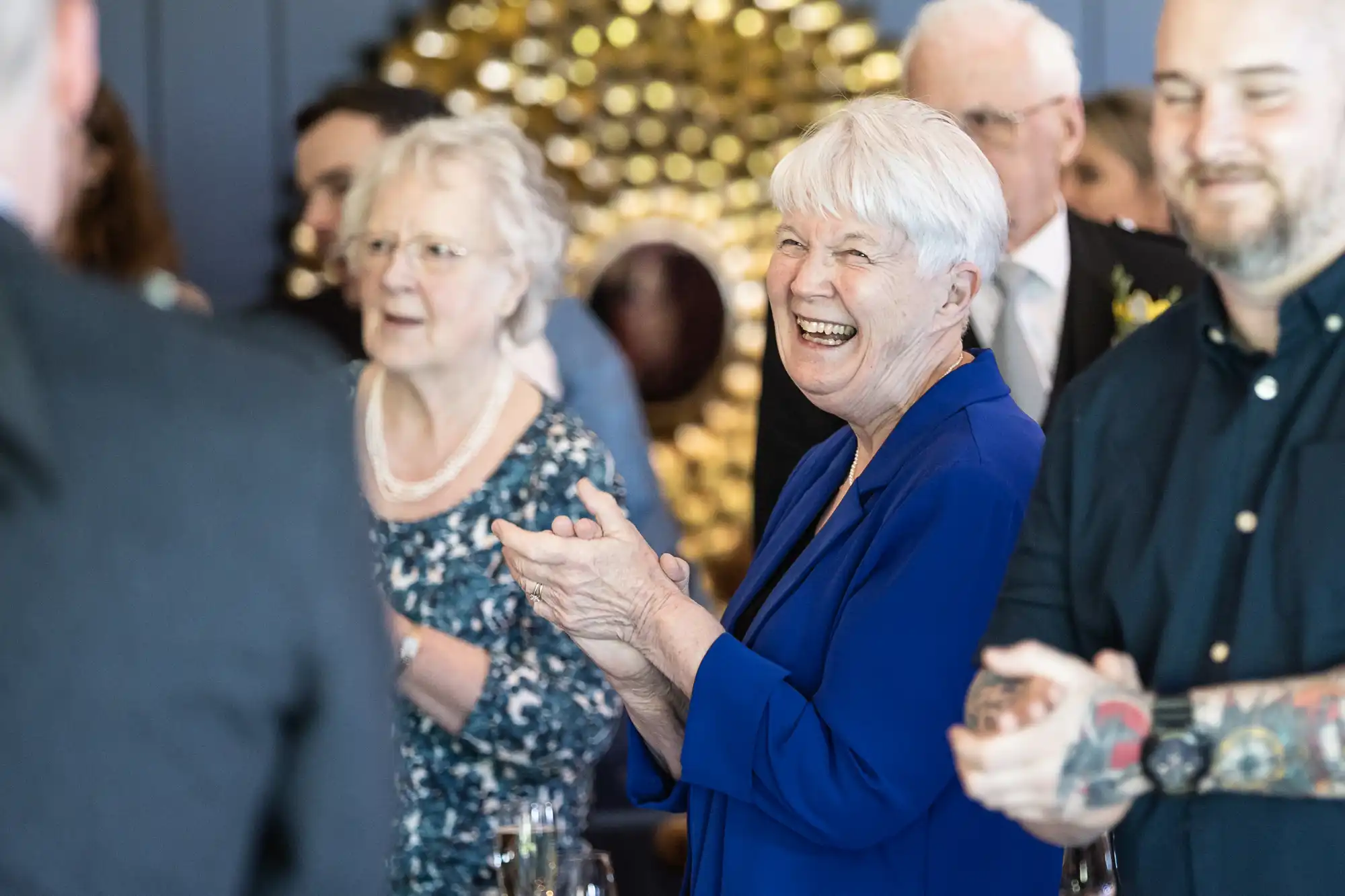 A group of people clapping and smiling at an event. An older woman in a blue jacket is at the center, appearing joyful. Others are partially visible in the background.