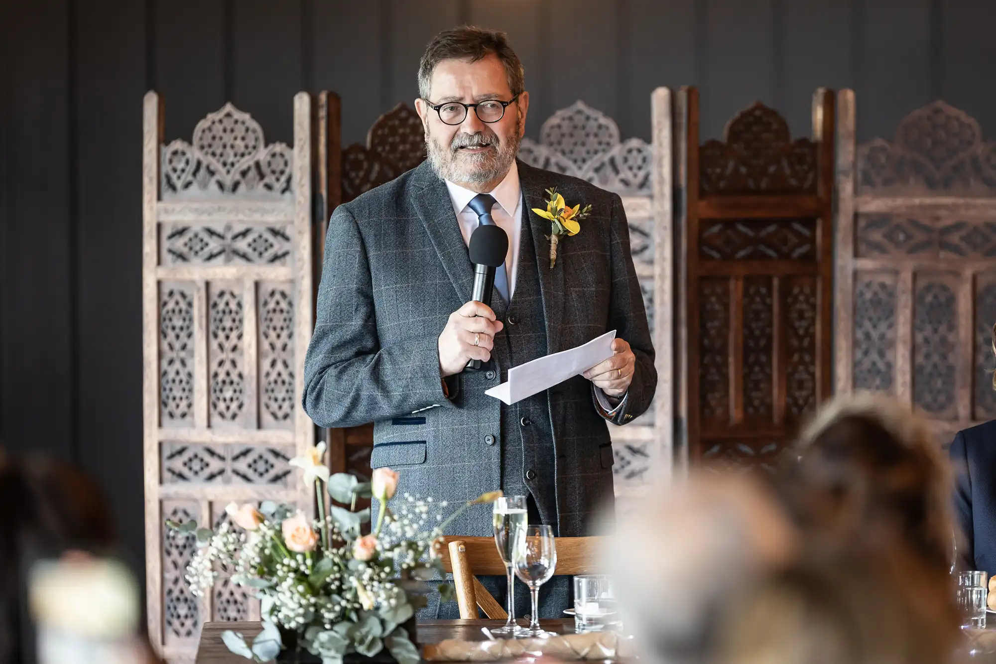 A man in a grey suit speaks into a microphone while holding a sheet of paper. Decorative screens and a floral arrangement are visible in the background.