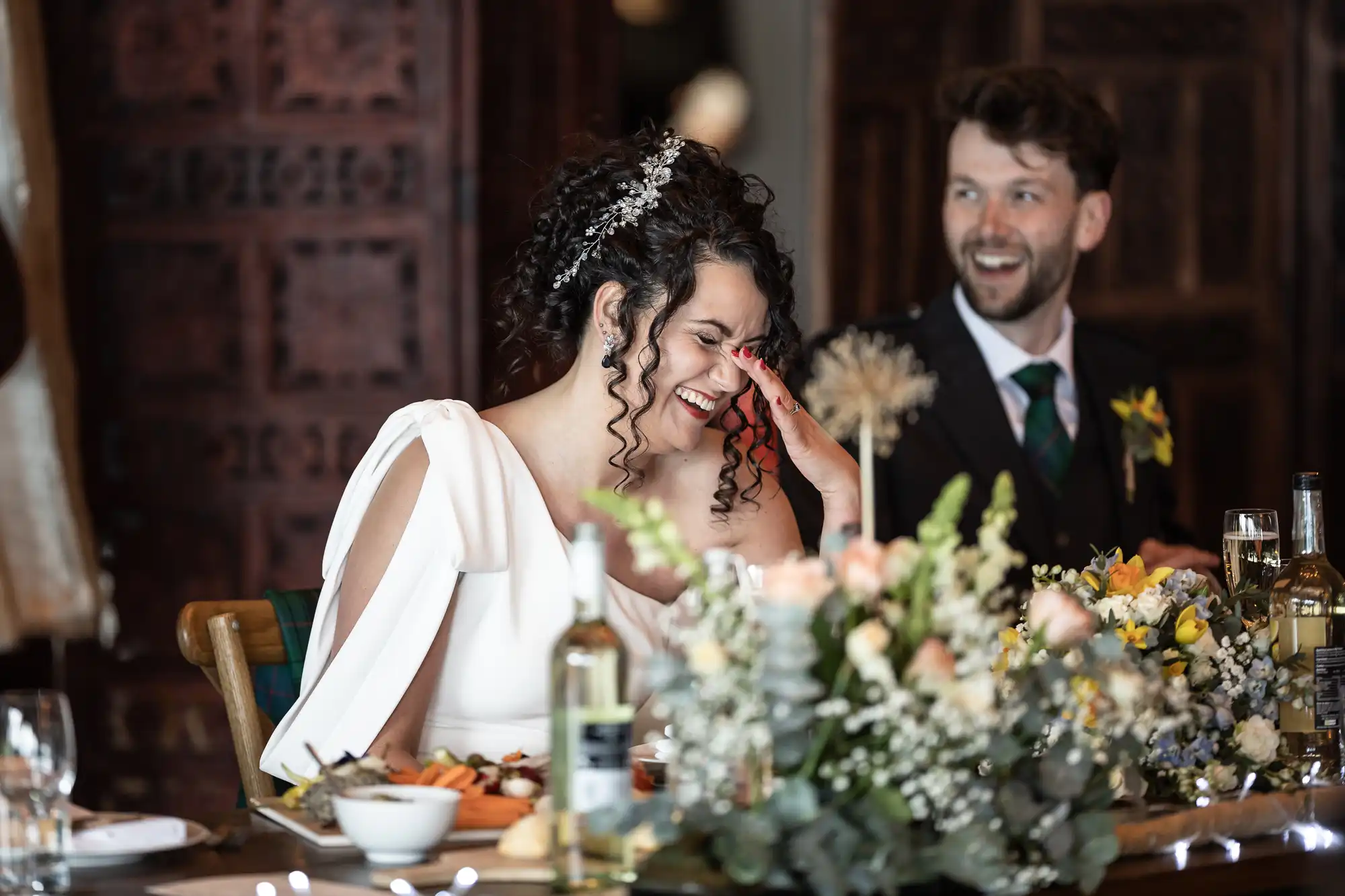 A bride and groom sit at a table adorned with flowers and wine, laughing and smiling during a wedding reception.