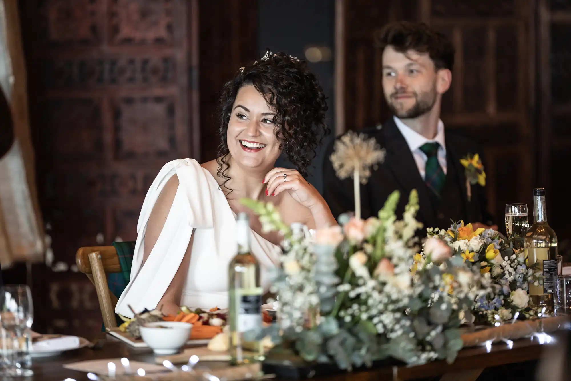 A bride in a white dress and a groom in a dark suit sit at a decorated table with food, drinks, and floral arrangements during a wedding reception.