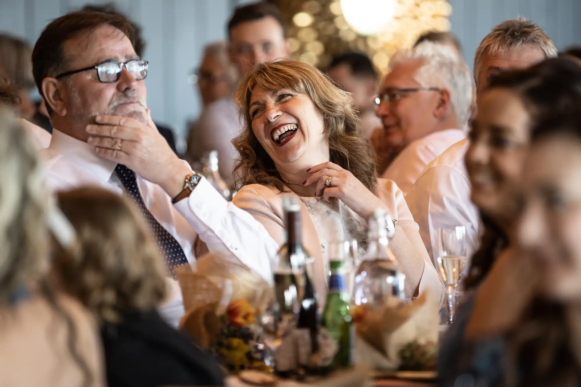 People seated at a table during a social event, with someone laughing heartily and others engaging in conversation.