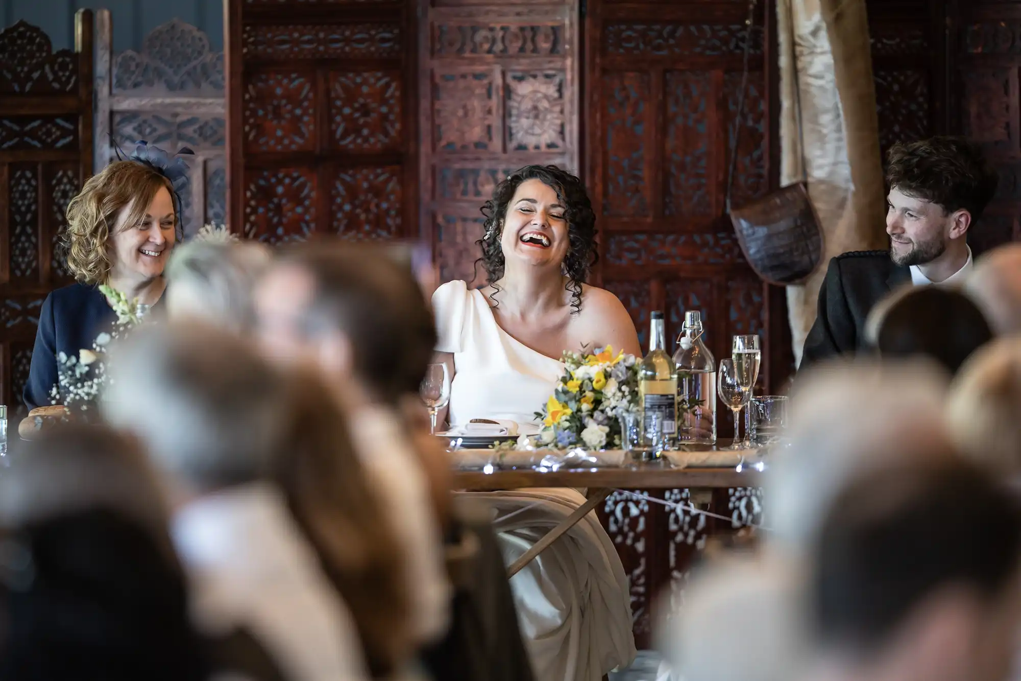 A smiling woman in a white dress sits at a table decorated with flowers and bottles, flanked by a man and another woman, in front of a wooden backdrop at a formal event.