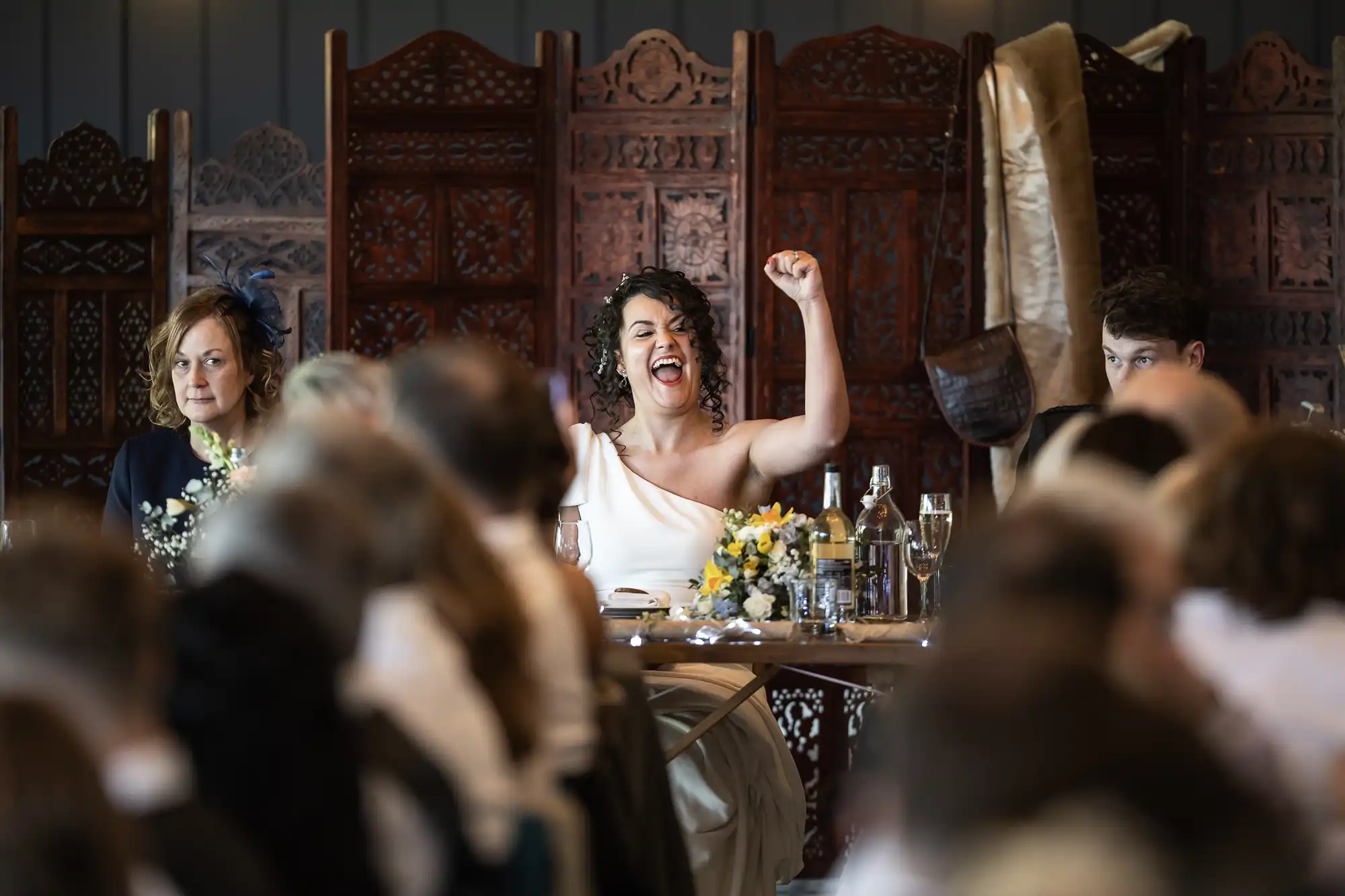 A person wearing a one-shoulder dress raises both arms in celebration or excitement while sitting at a table in front of ornately carved wooden screens. Other people are seated at the same table.