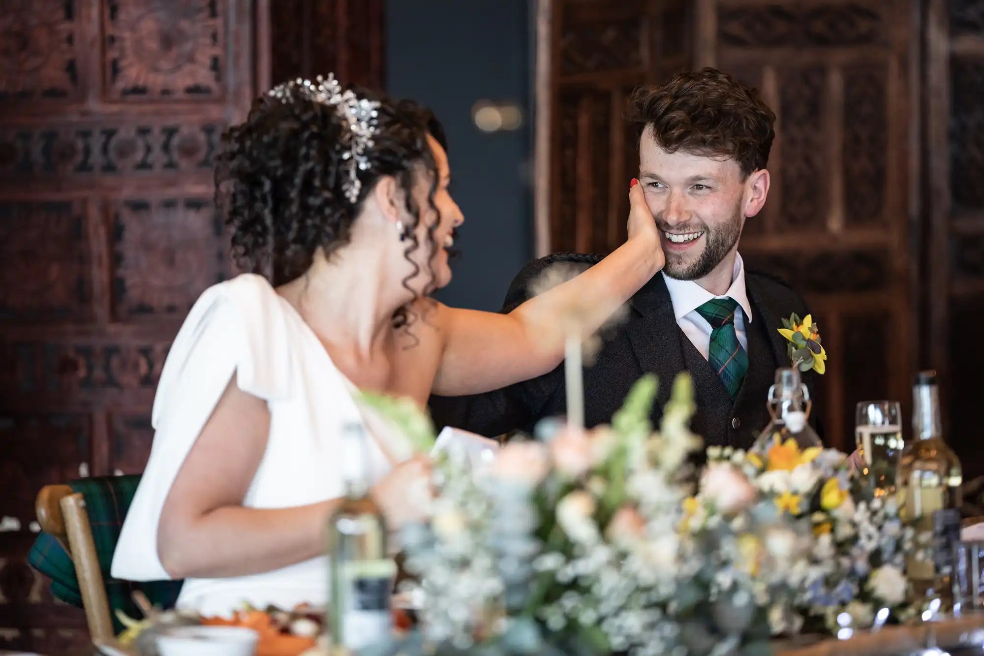 A bride in a white dress and a groom in a dark suit with a yellow boutonniere share a joyful moment at a wedding reception table adorned with flowers.