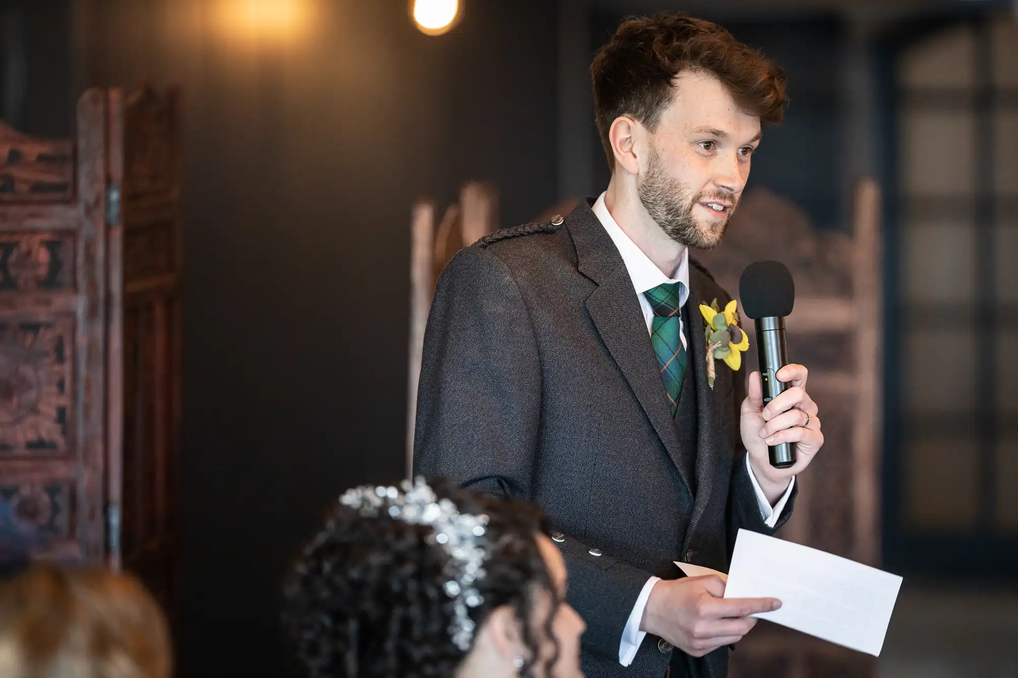 A man wearing a suit speaks into a microphone while holding a piece of paper. He is indoors with blurred background elements behind him.