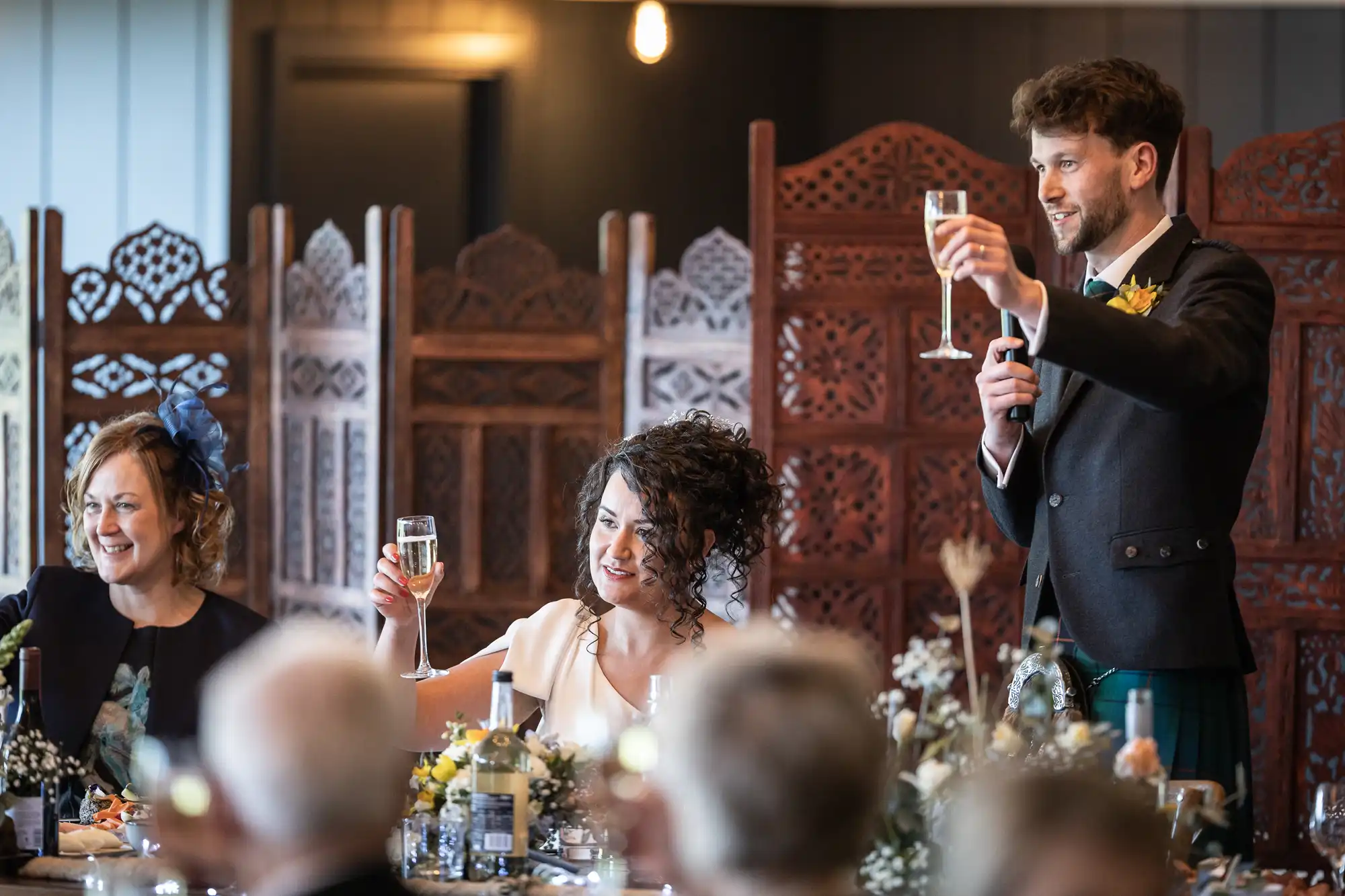 Three people at a celebration raise their glasses for a toast. The man is standing and smiling, holding a microphone in one hand and a glass of champagne in the other.