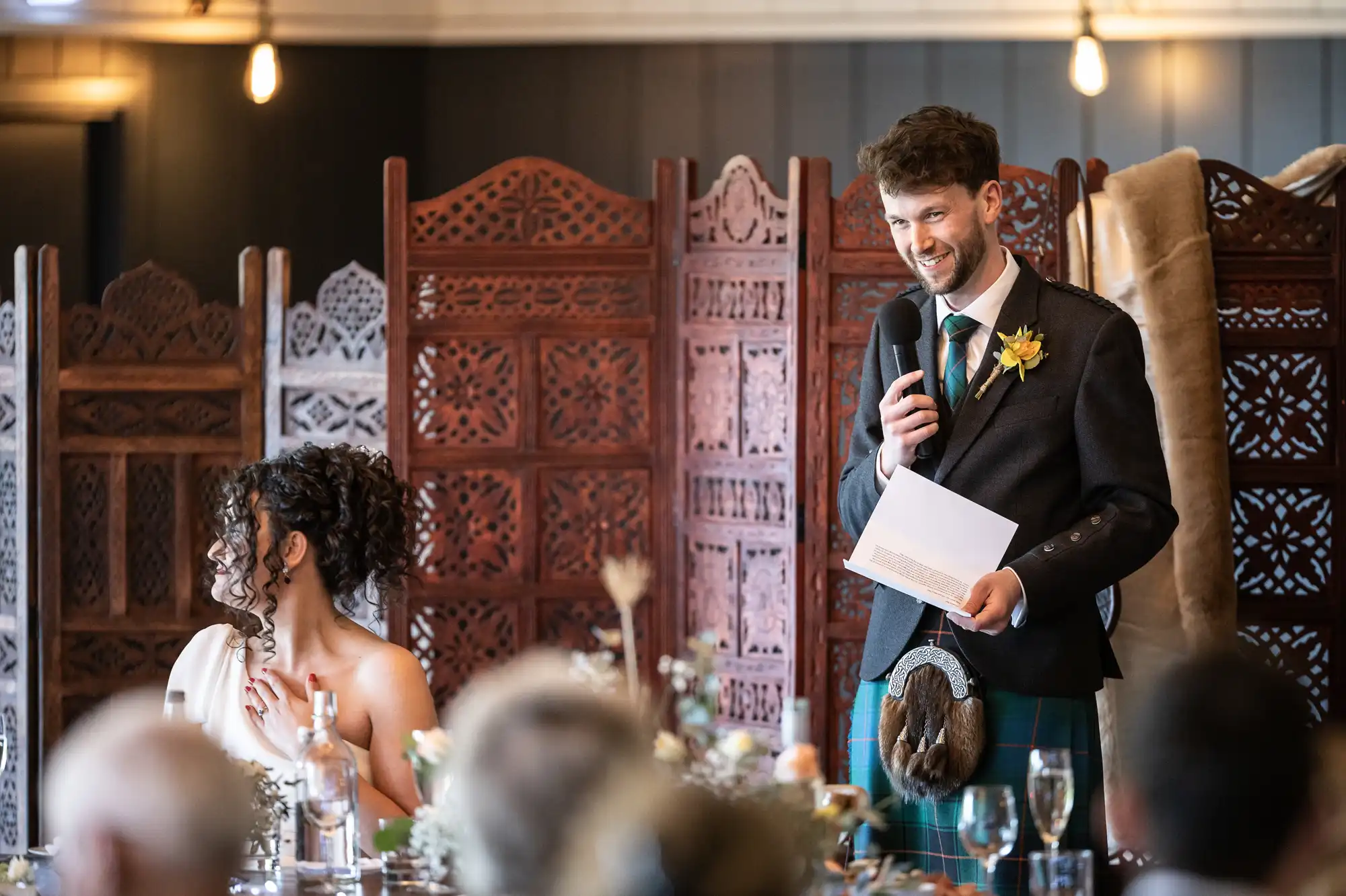 A man in formal attire speaks into a microphone while holding a sheet of paper. A woman in a white dress sits beside him, listening. They are in a decorated room with wooden screens in the background.