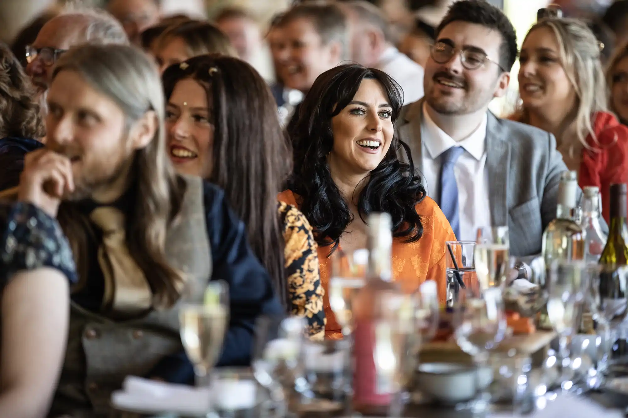 A group of people, dressed in formal attire, are sitting at a table and smiling while watching something off-camera at an event. Various drinks and glassware are on the table in front of them.