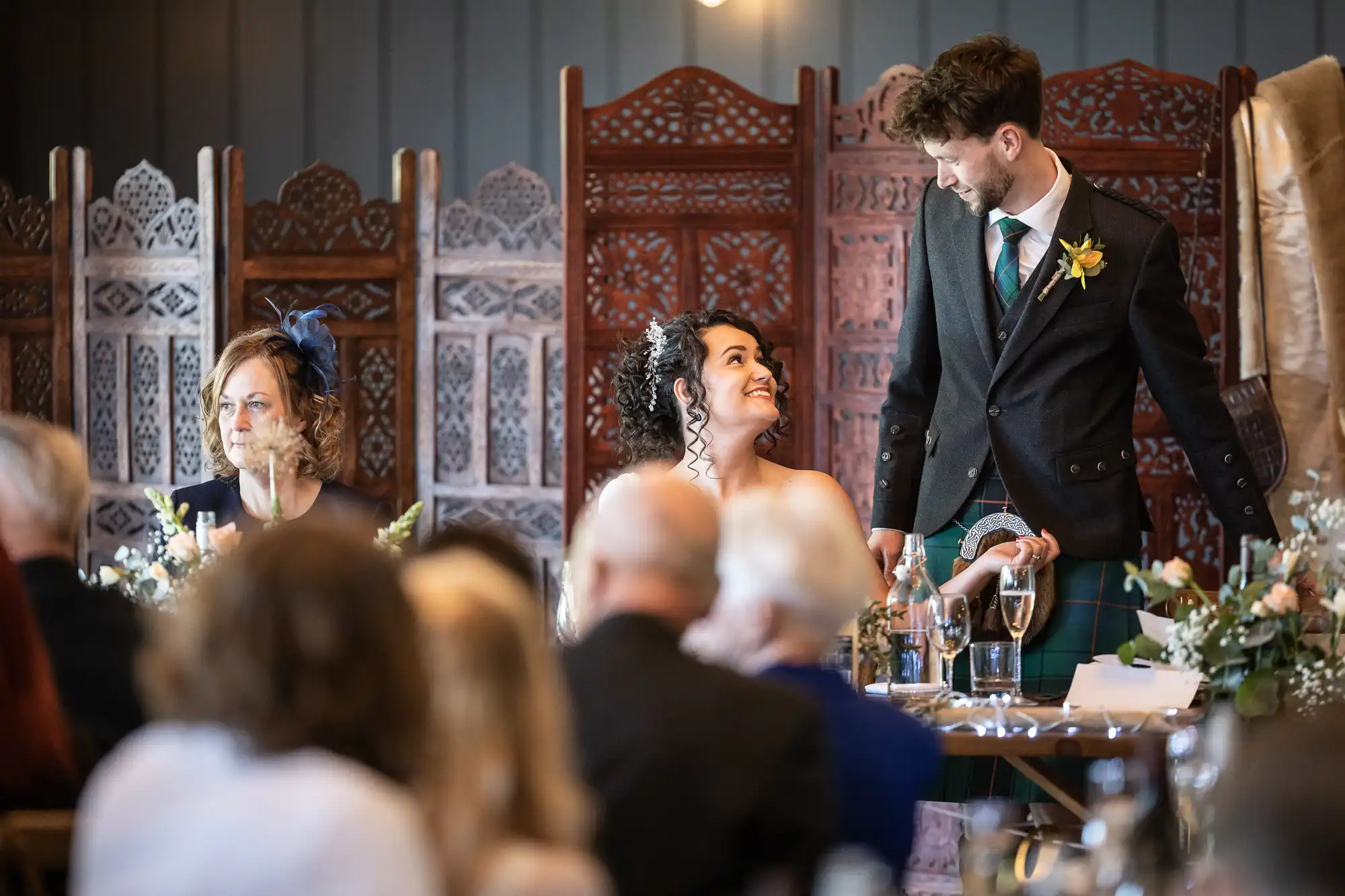 A person with curly hair in a white dress and a person in a suit smile and hold hands at a formal event, while another person with a fascinator sits beside them at a table with floral arrangements.