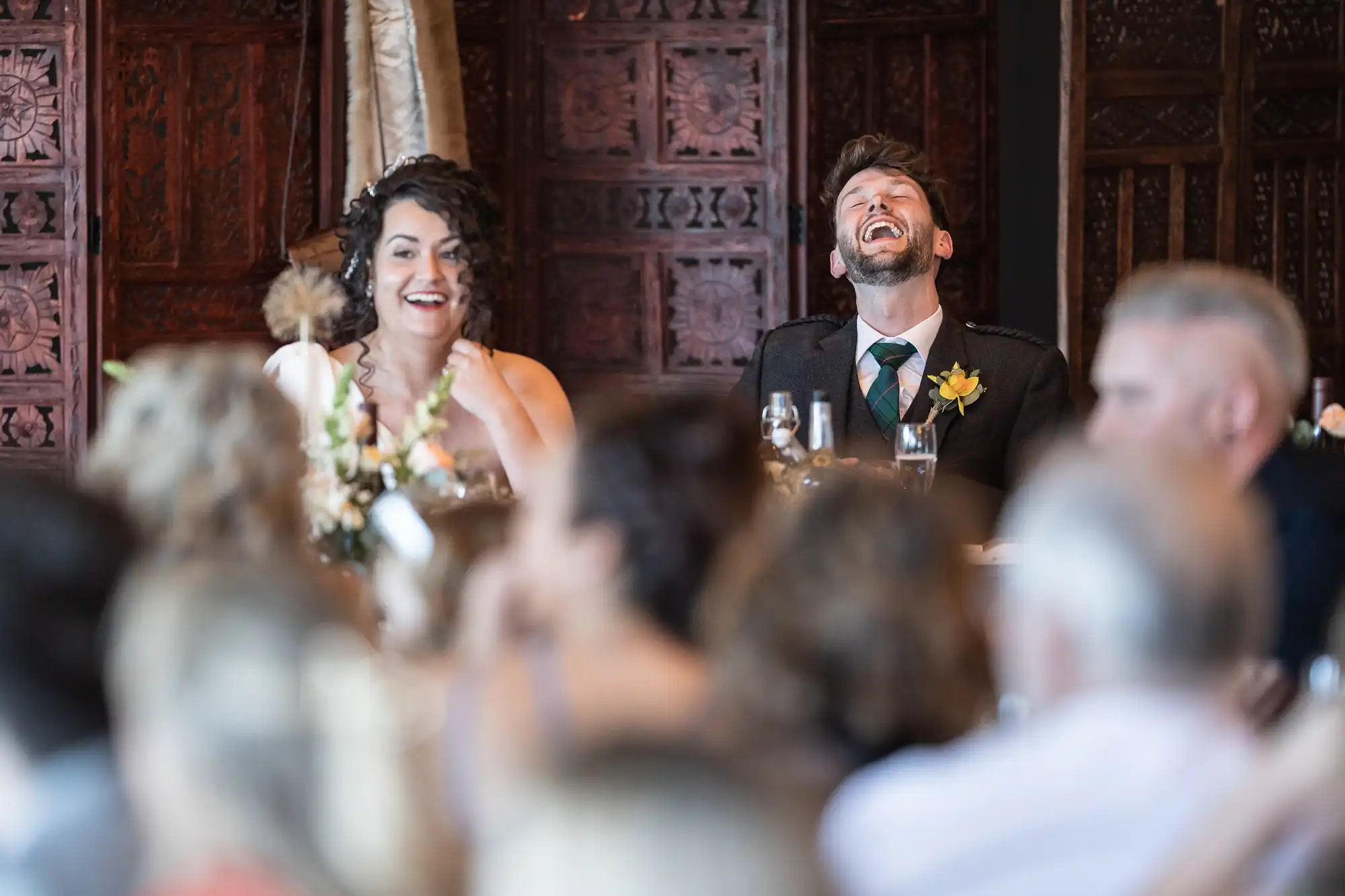 Two people, dressed formally, sit at a table, laughing heartily during an indoor event with several attendees.