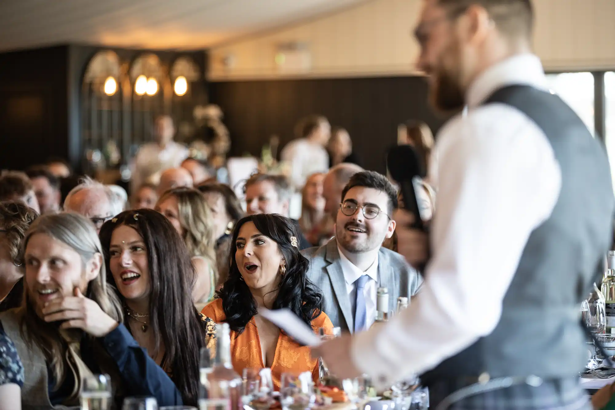 A group of people seated at dining tables look towards a standing person giving a speech in a room with a festive atmosphere.