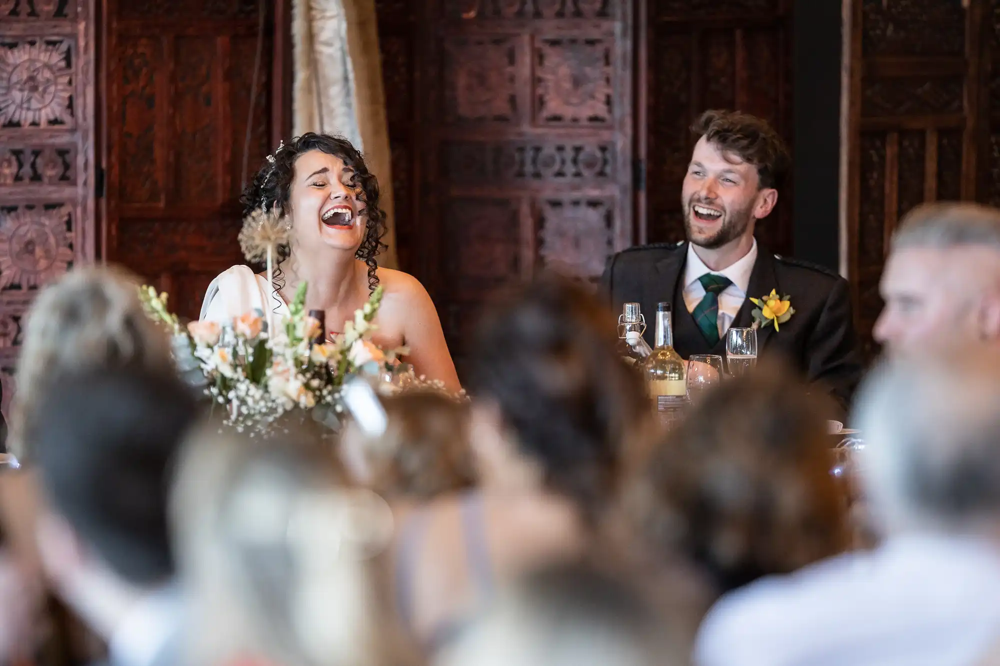 A couple is sitting at a table during a wedding reception, smiling and laughing. The background features ornate wooden carvings, and there are floral arrangements and bottles on the table.