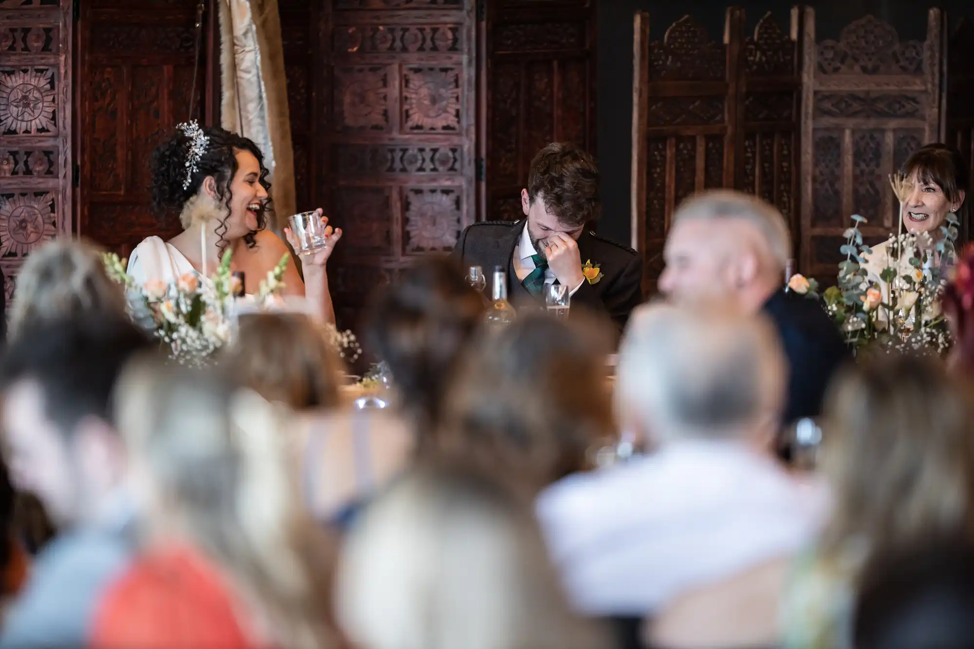 People are seated at a wedding reception. A bride laughs while the groom covers his face with his hand. Guests are seated around them, with the background decorated with ornate wooden panels.