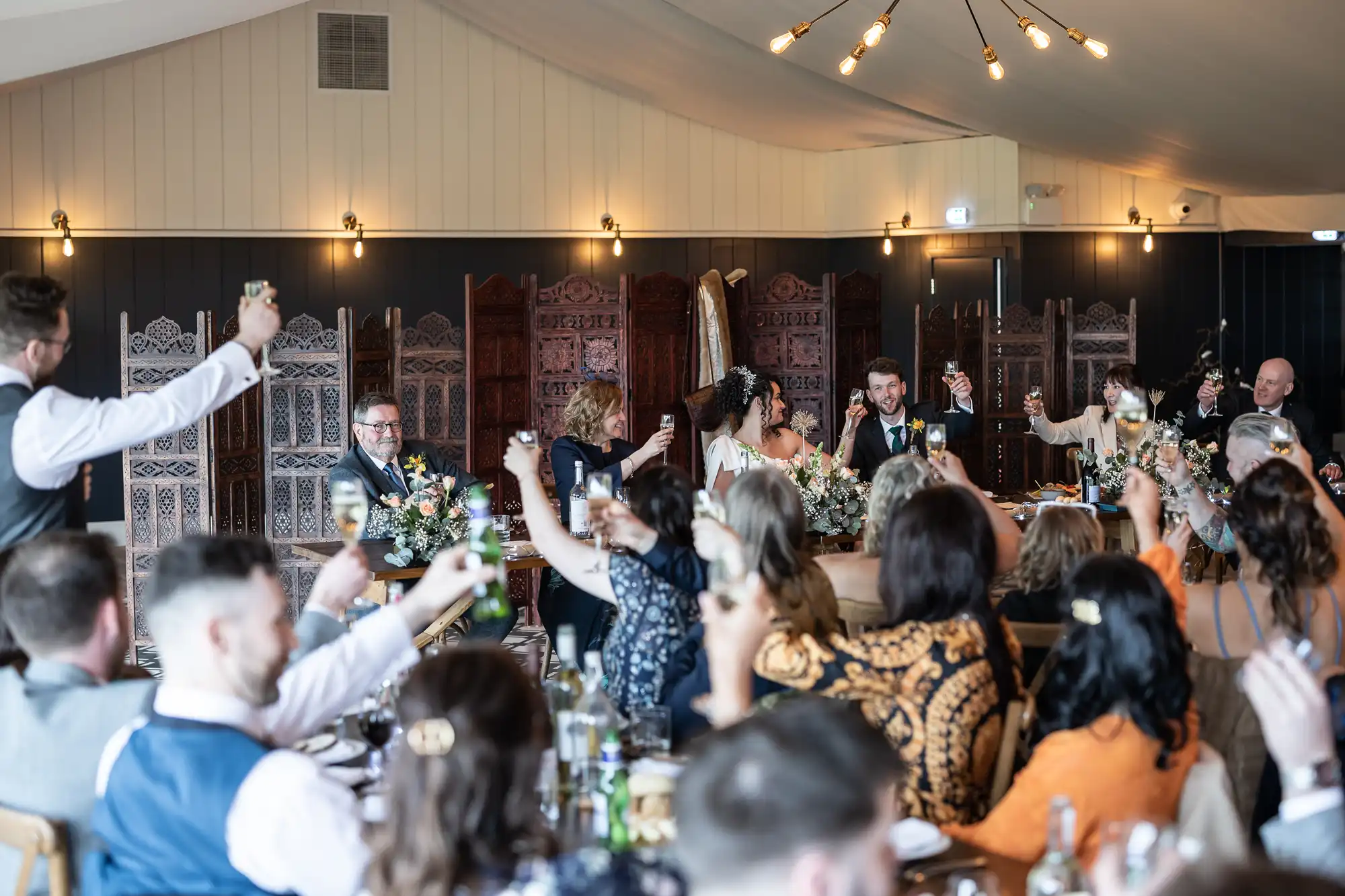 A group of people seated at a long table, raising glasses in a toast in a warmly lit room with ornate wooden partitions in the background.